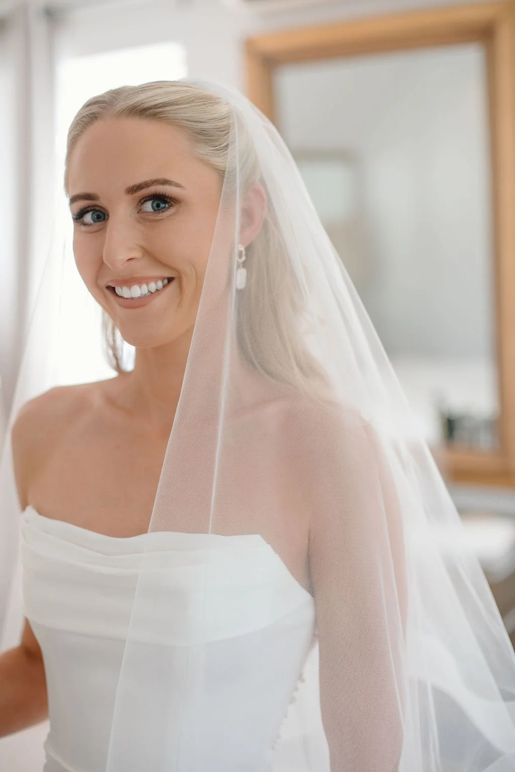 Bride with blonde hair in a wedding dress and veil, smiling, indoors with mirror and window in background.Bride with a fresh, natural bridal makeup look and softly styled hair.