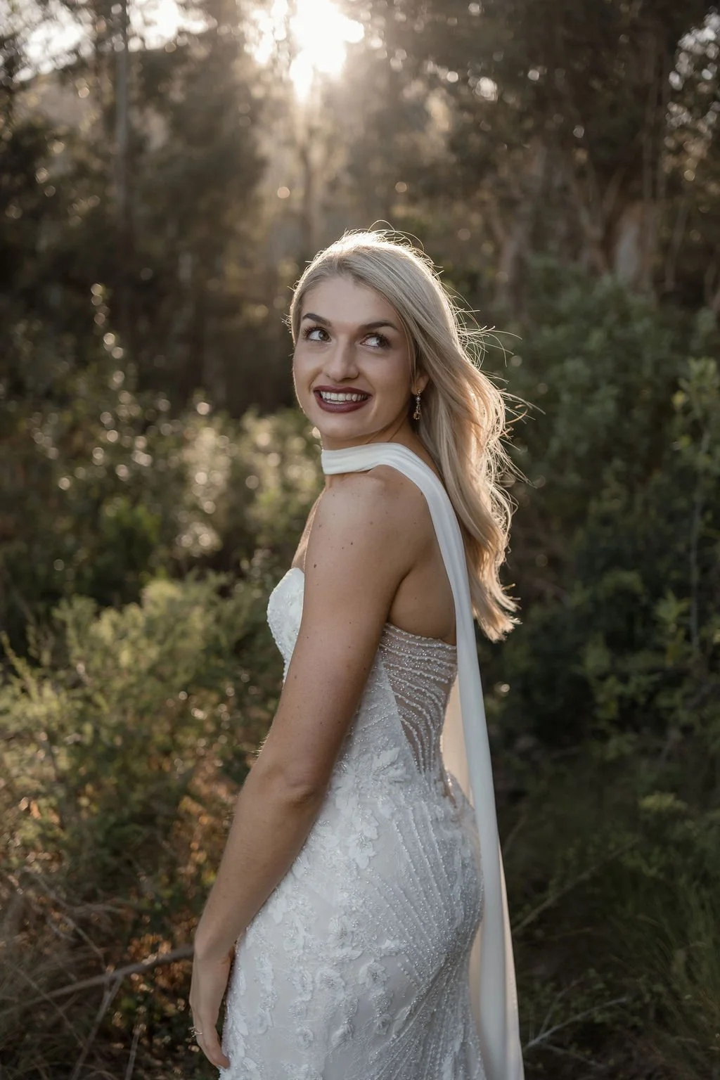 A smiling blonde woman in a white wedding dress with intricate beading, standing outdoors among trees as the sun sets in the background.