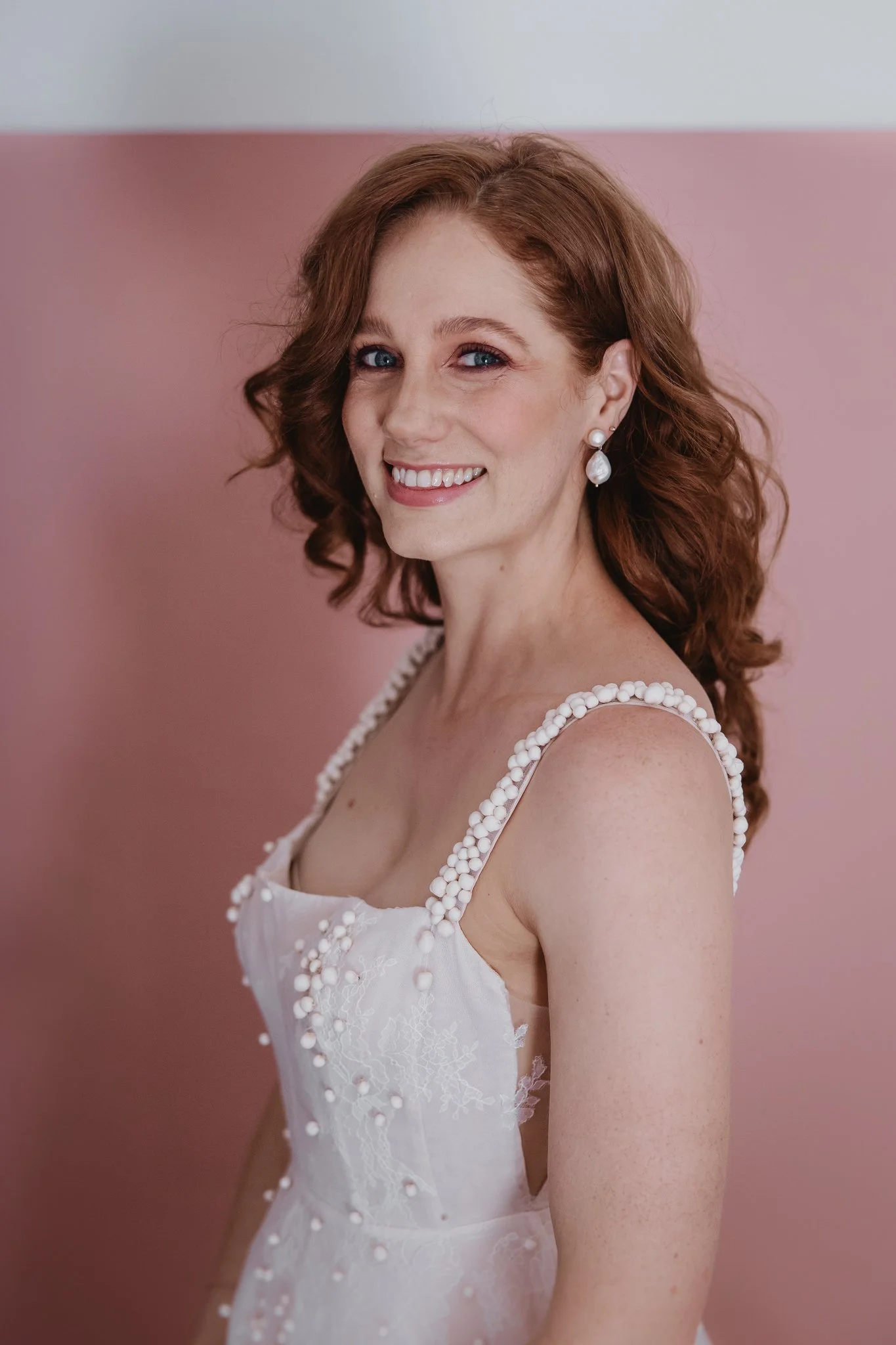 A woman with red hair, blue eyes, and fair skin wearing pearl earrings and a white dress with pearl straps, standing against a pink and gray background, smiling.