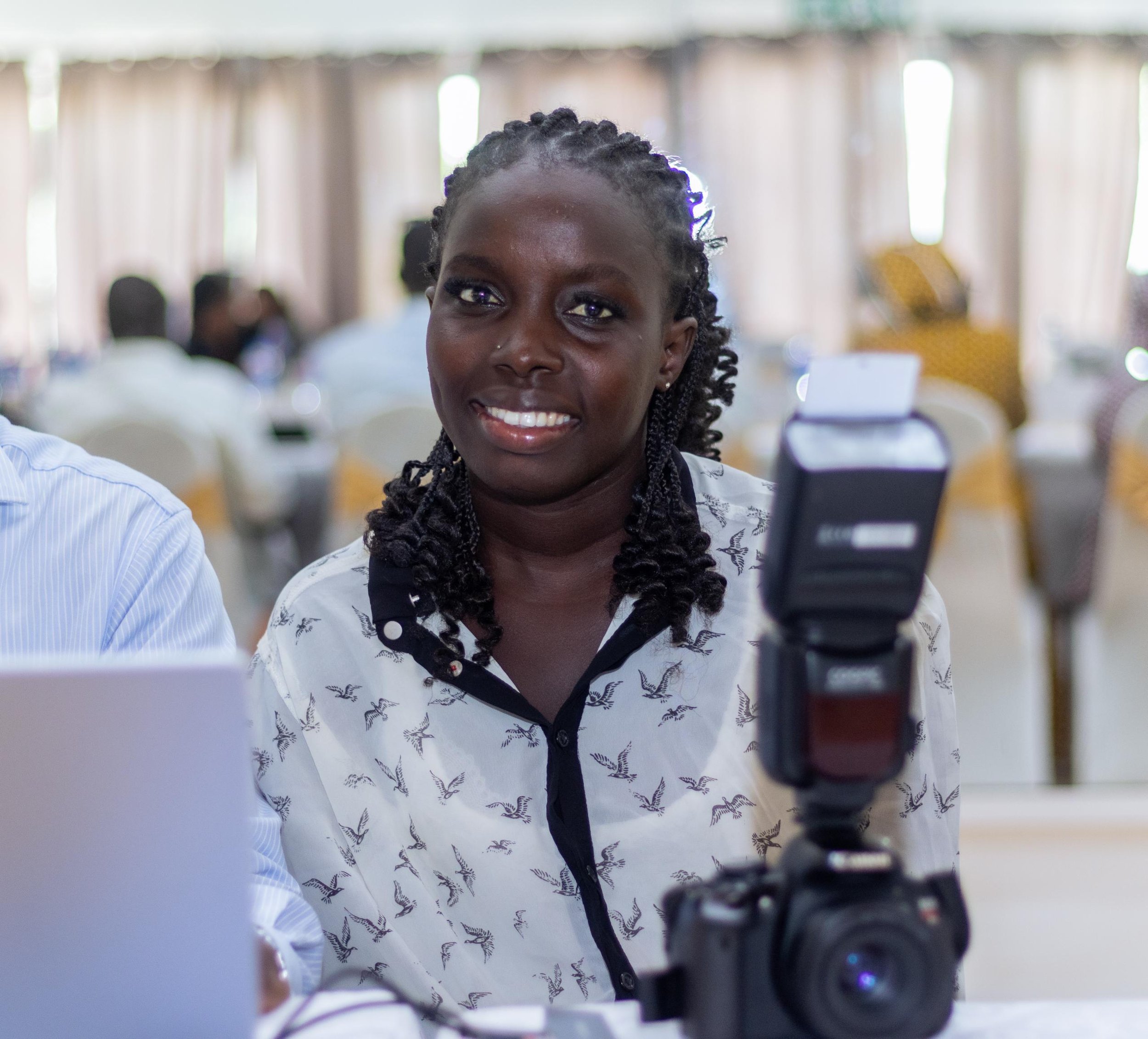 A smiling woman sitting at a table with a camera in front of her, in a bright indoor setting with other people in the background.