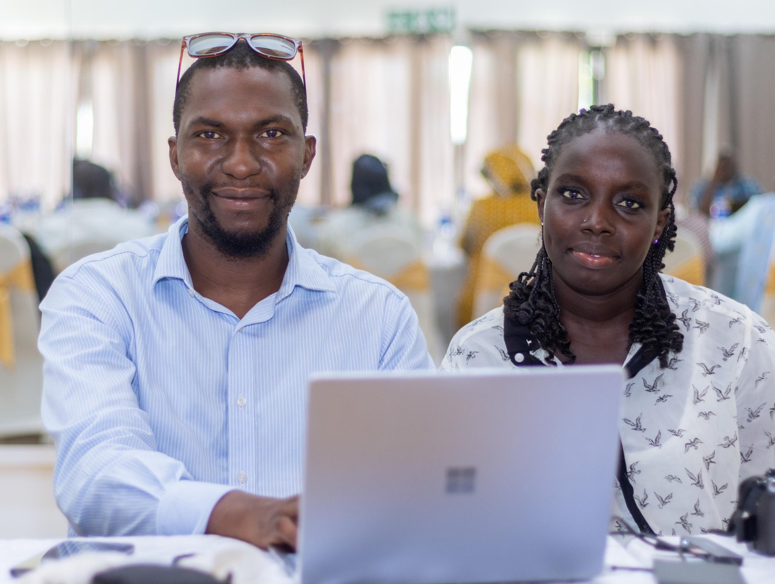 A man and woman sitting at a table with a silver laptop in front of them in a bright room with many windows and other people in the background.