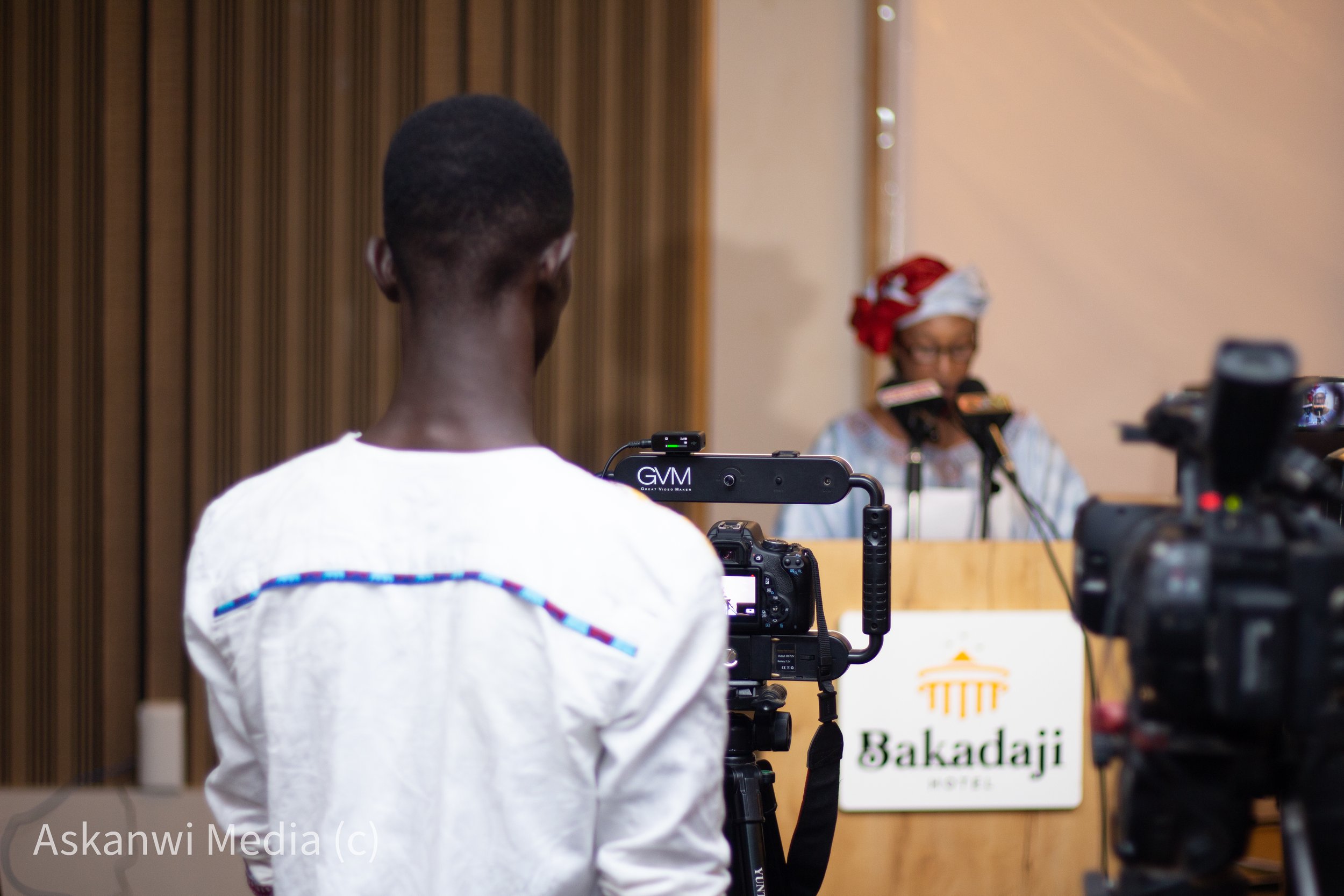 A woman wearing a head wrap speaking at a podium with a sign that reads "Bakamdji" at a conference, with a videographer filming her.