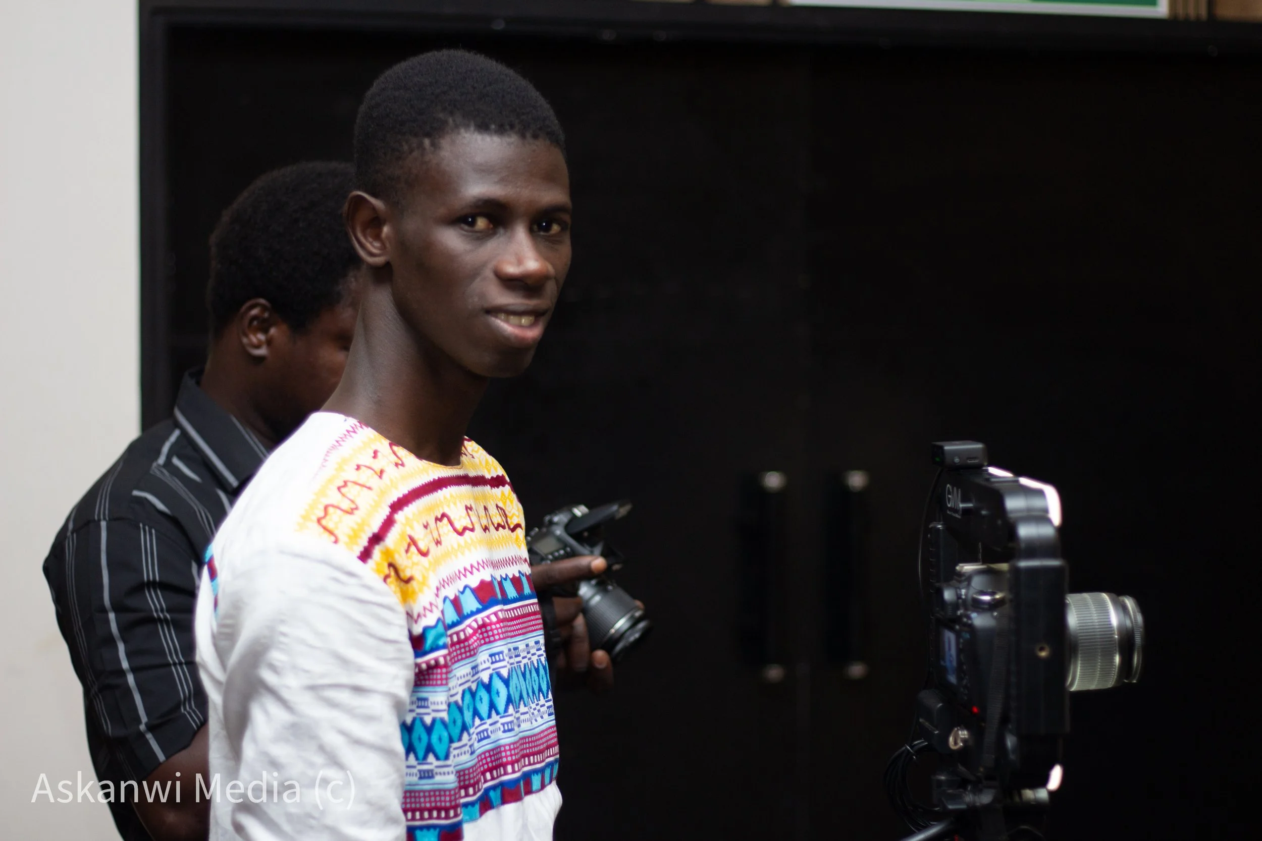 Young man in colorful traditional clothing stands near camera equipment, with another person behind him holding a camera, in a dark room.