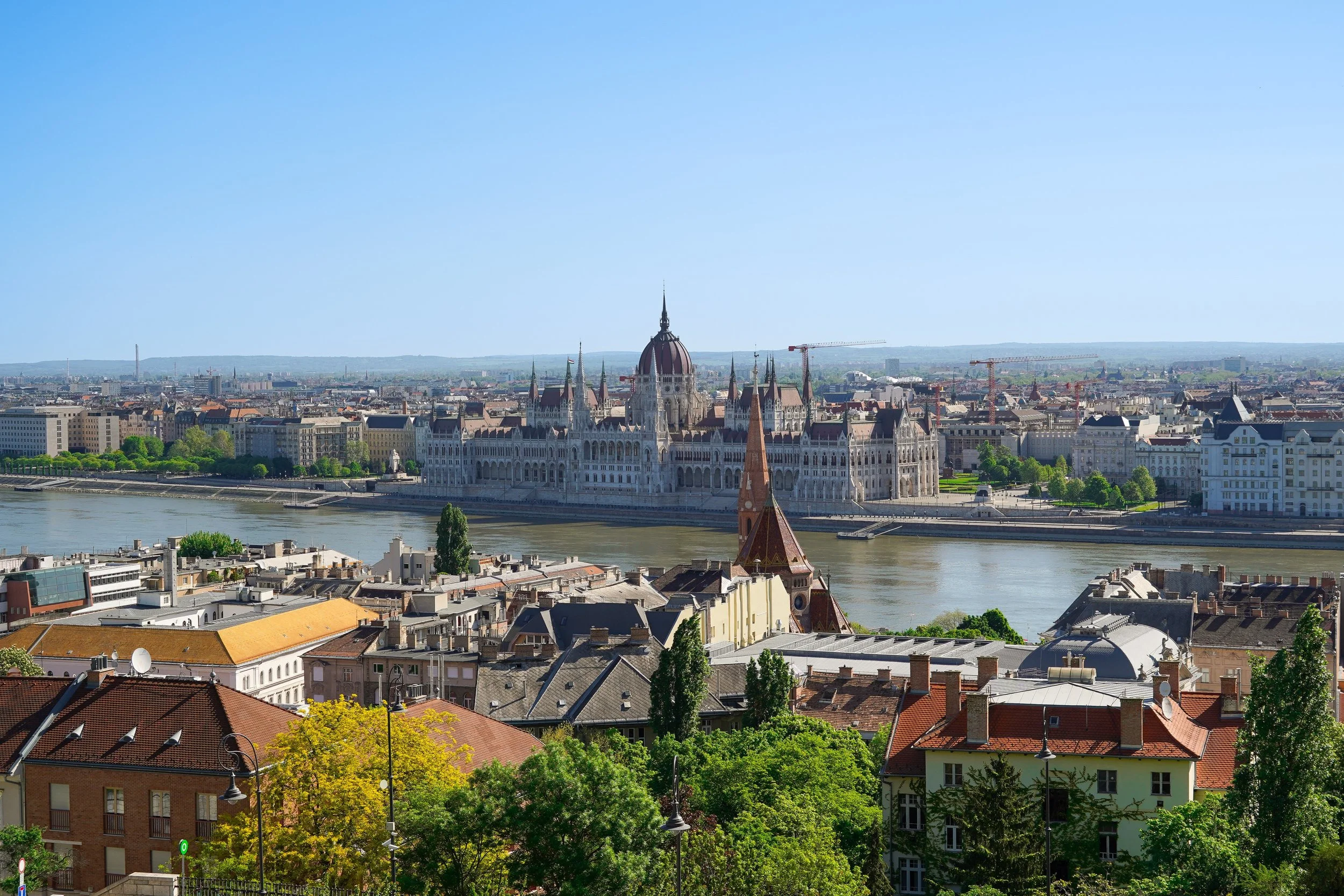 A panoramic cityscape of Budapest, Hungary, showing the Parliament building on the banks of the Danube River, with historic rooftops and green trees in the foreground. Photo by Carla Hugues-Moscariello, Photographer & Travel Content Creator