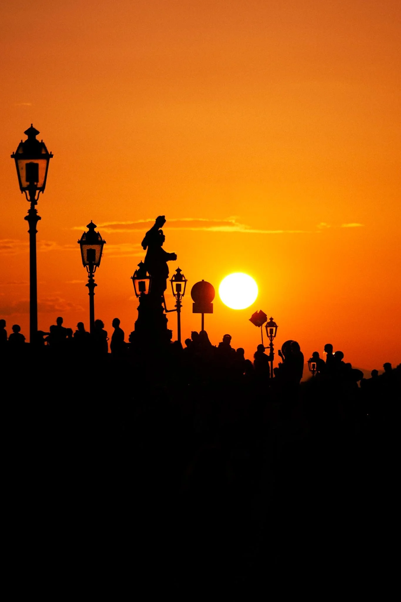Silhouetted people and vintage street lamps at sunset with a bright sun in the sky.