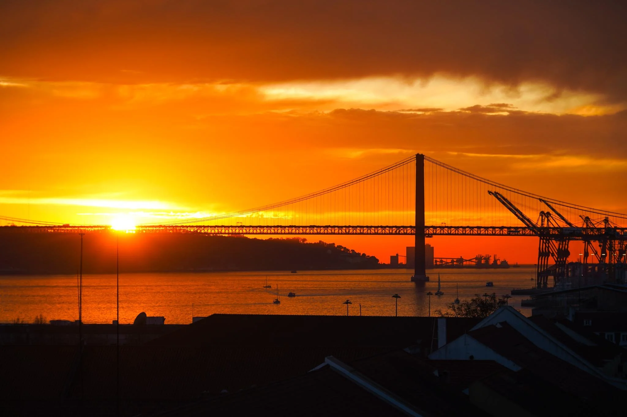 Sunset over a bridge with a dark sky and orange clouds, with boats on the water and cranes on the right.