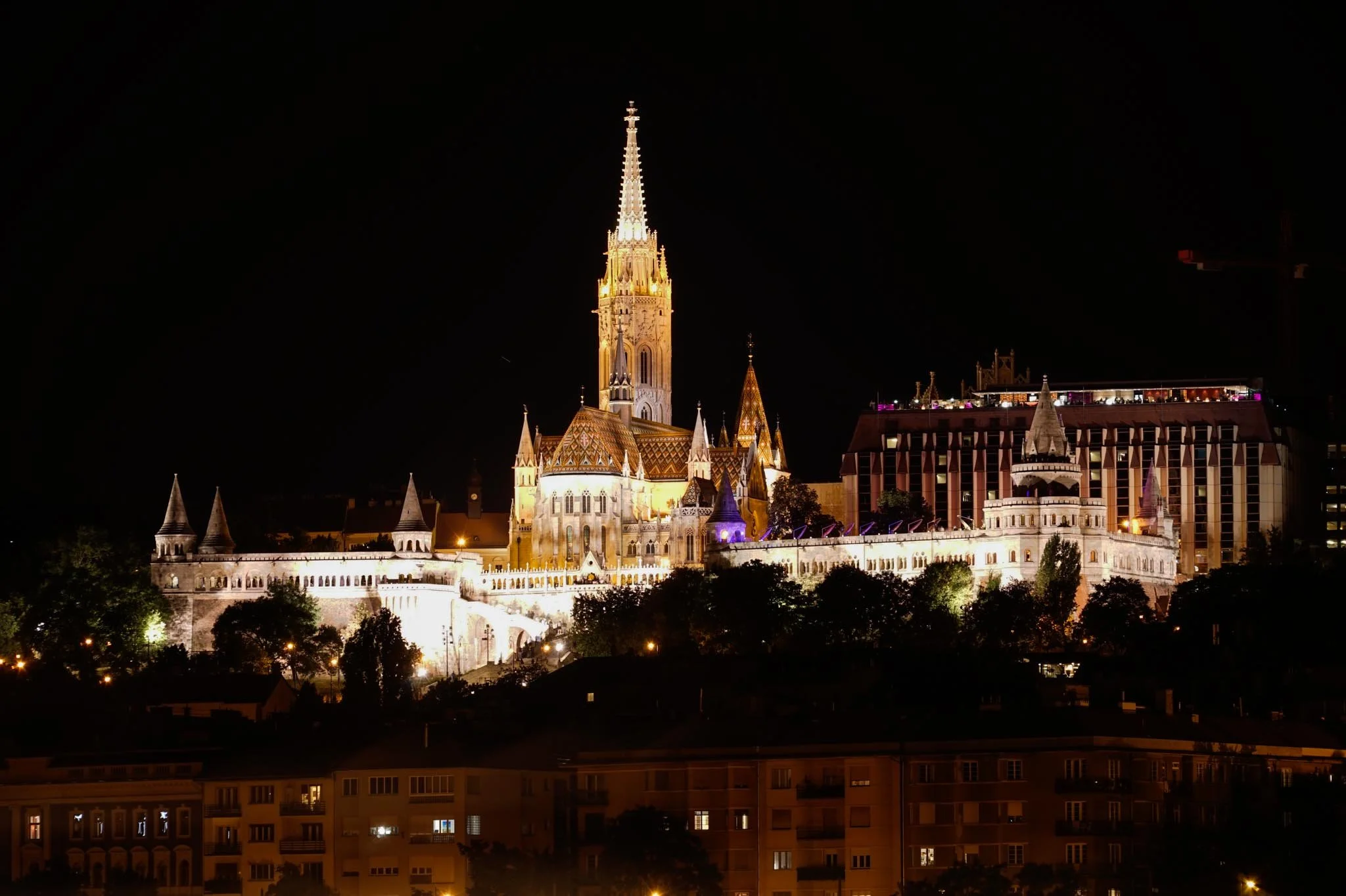 Night view of a brightly illuminated castle with a tall, pointed spire, surrounded by smaller towers and buildings, set against a dark night sky, with some trees and buildings in the foreground.