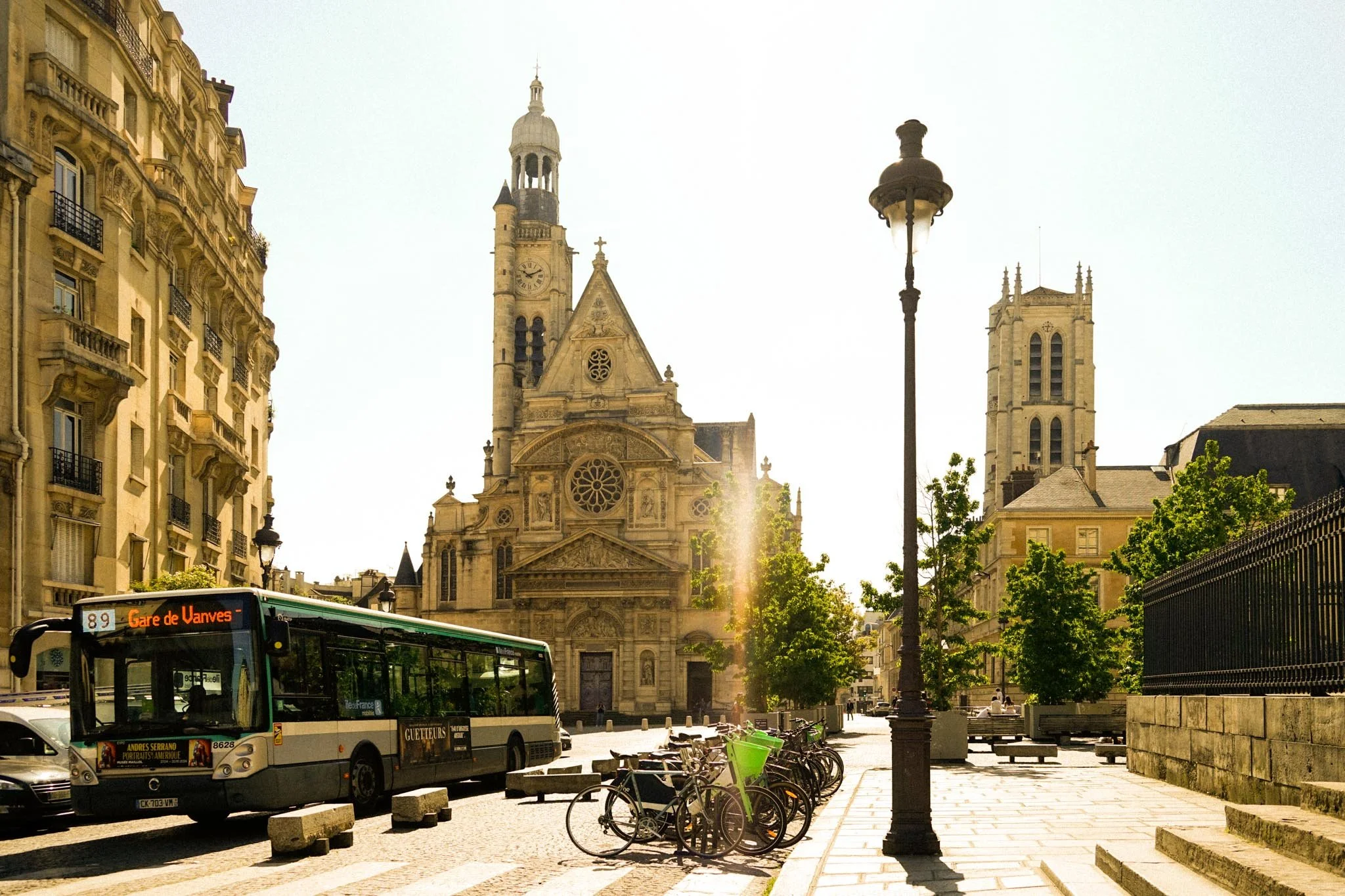 A street view in front of a historic church with a bus, bicycles, trees, benches, and a lamppost.