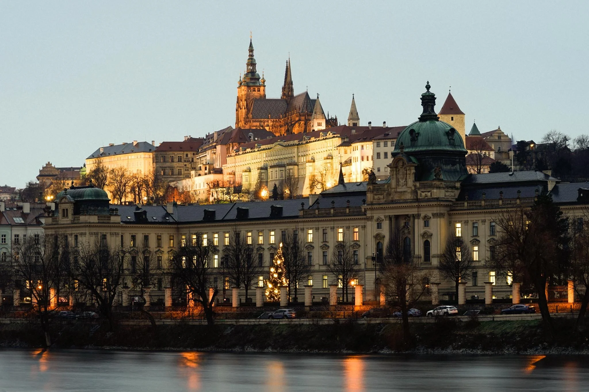 A cityscape view of Prague at dusk with illuminated historic buildings and a church on a hill, near a riverfront with trees and a decorated Christmas tree.