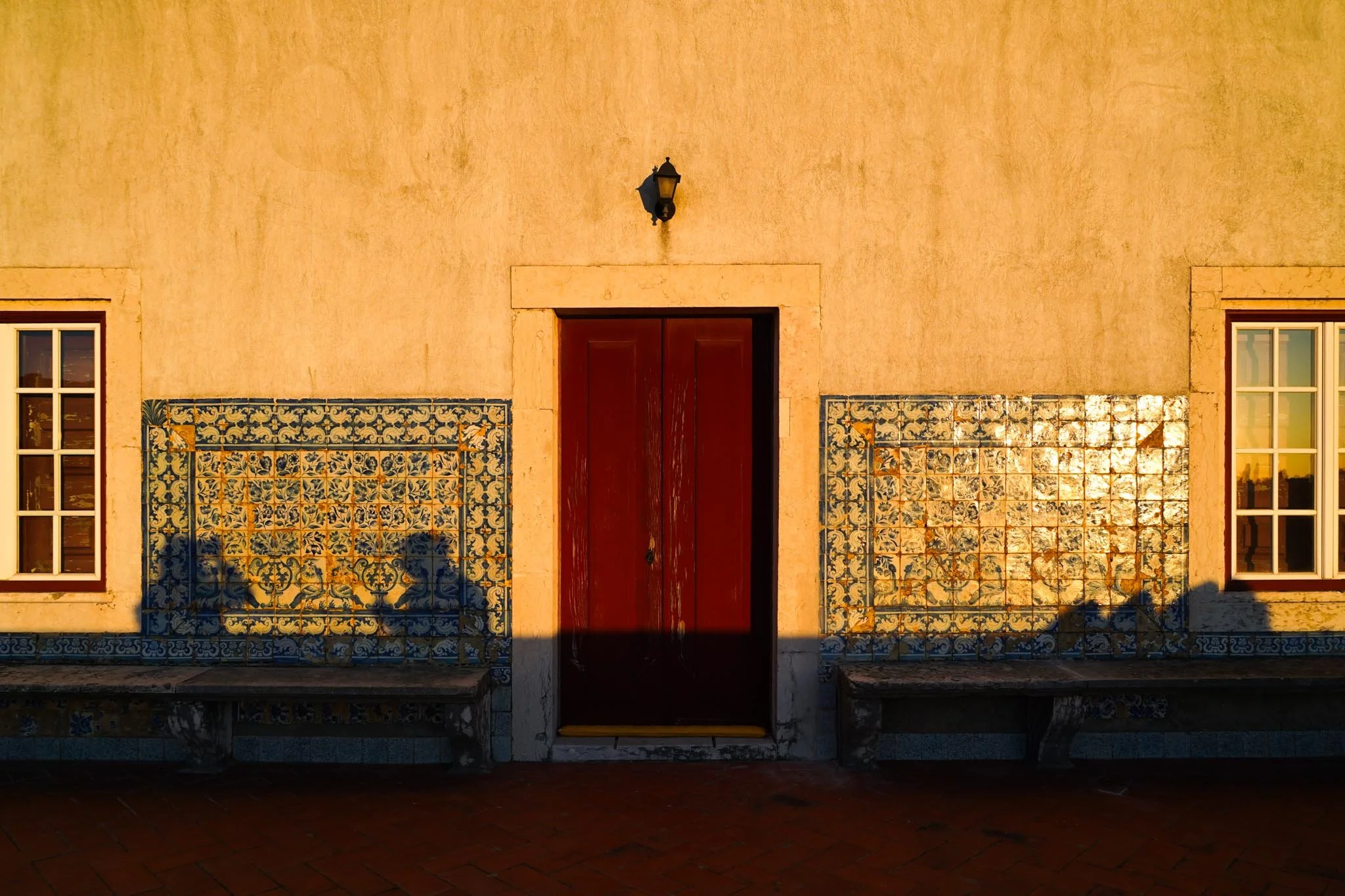 A beige building wall with two windows and a red wooden door in the center. The wall is decorated with blue and yellow patterned tiles on either side of the door, and there are two benches below the tiles. The shadow of a candelabrum is cast onto the