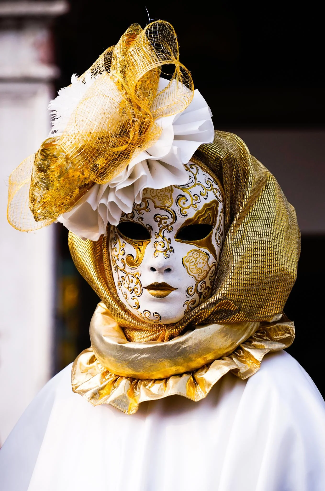 Person wearing a white mask decorated with gold patterns, black eye holes, and gold lips, dressed in gold and white garments, with gold and white headpieces and accessories.