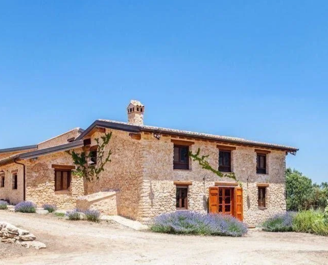 Casa de piedra con puertas de madera y plantas en el jardín, bajo cielo azul.