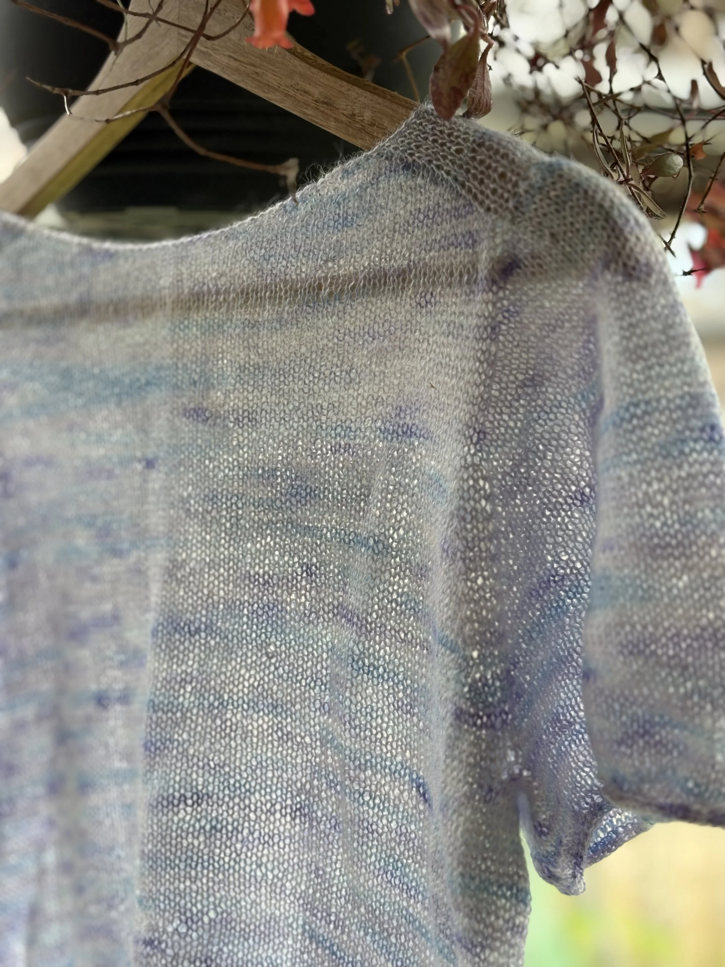 Close-up of a sheer, iridescent fabric shirt hanging on a wooden hanger with flowers and greenery in the background.