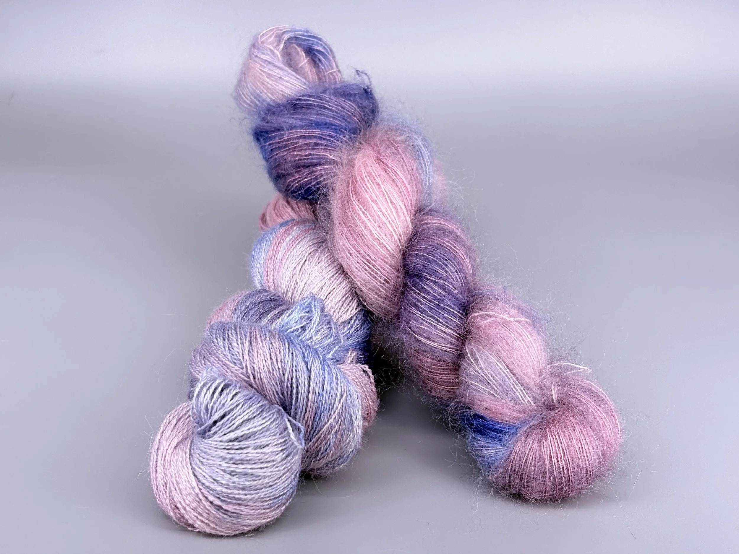 Multiple skeins of pastel-colored yarn in shades of pink, purple, and blue, twisted into a loose bundle against a plain, light background.