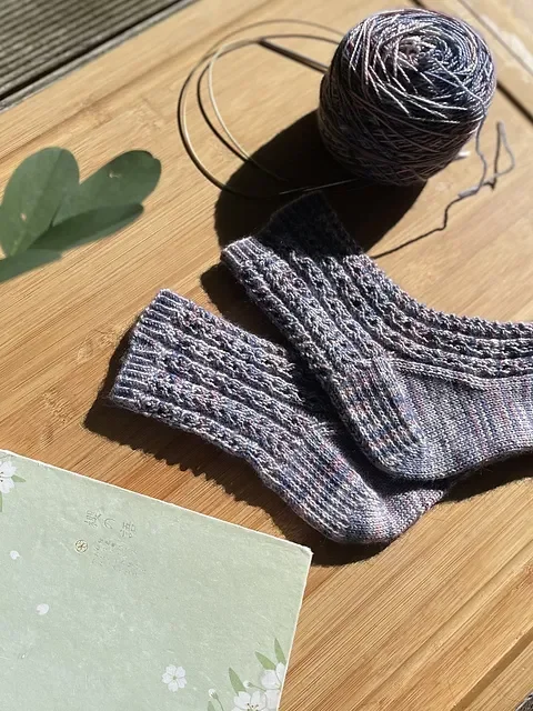 Gray knit socks, a ball of yarn, and a leaf on a wooden surface.