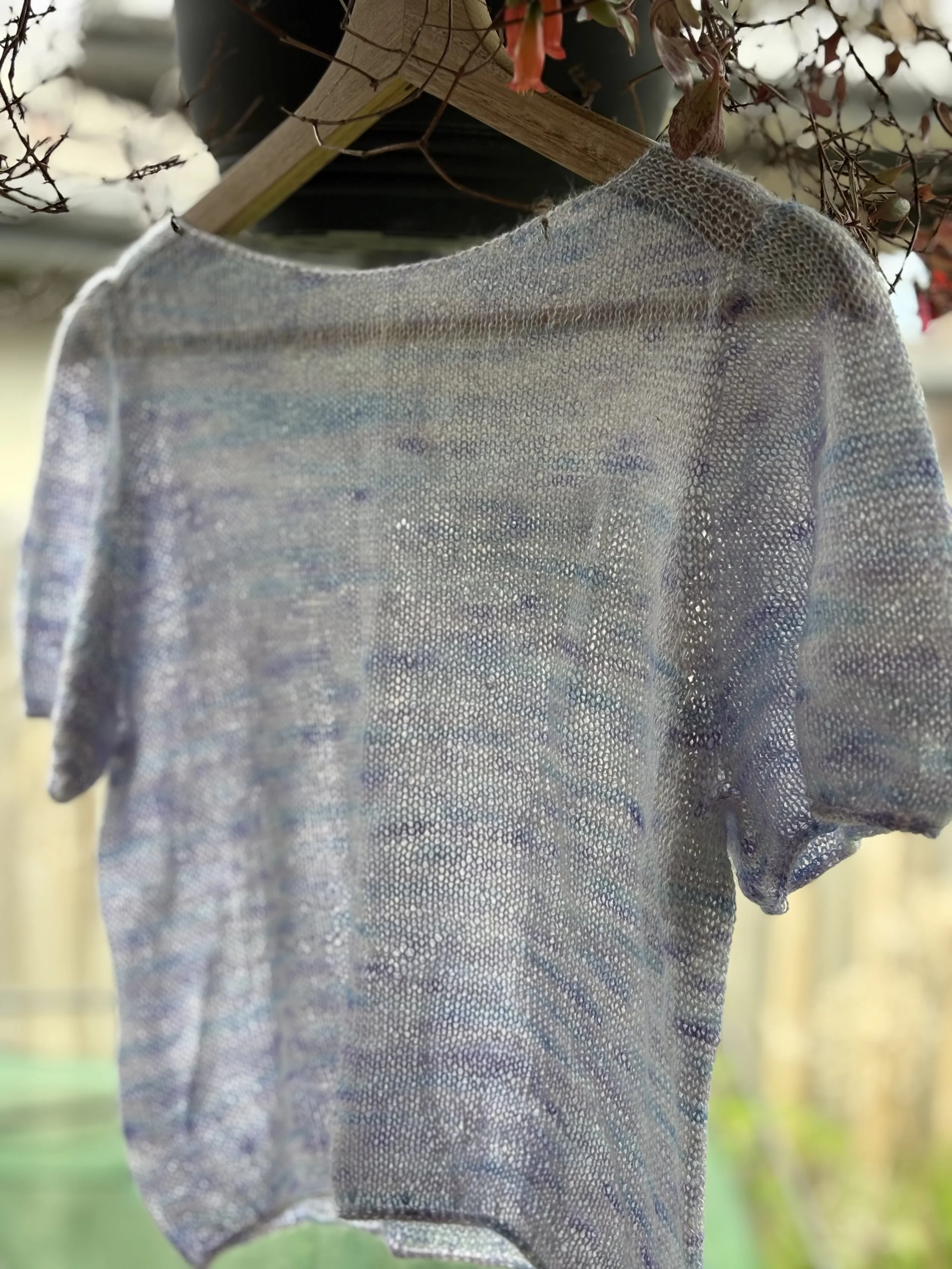 A sheer, knitted, multicolored blouse with short sleeves hanging on a wooden hanger outdoors.