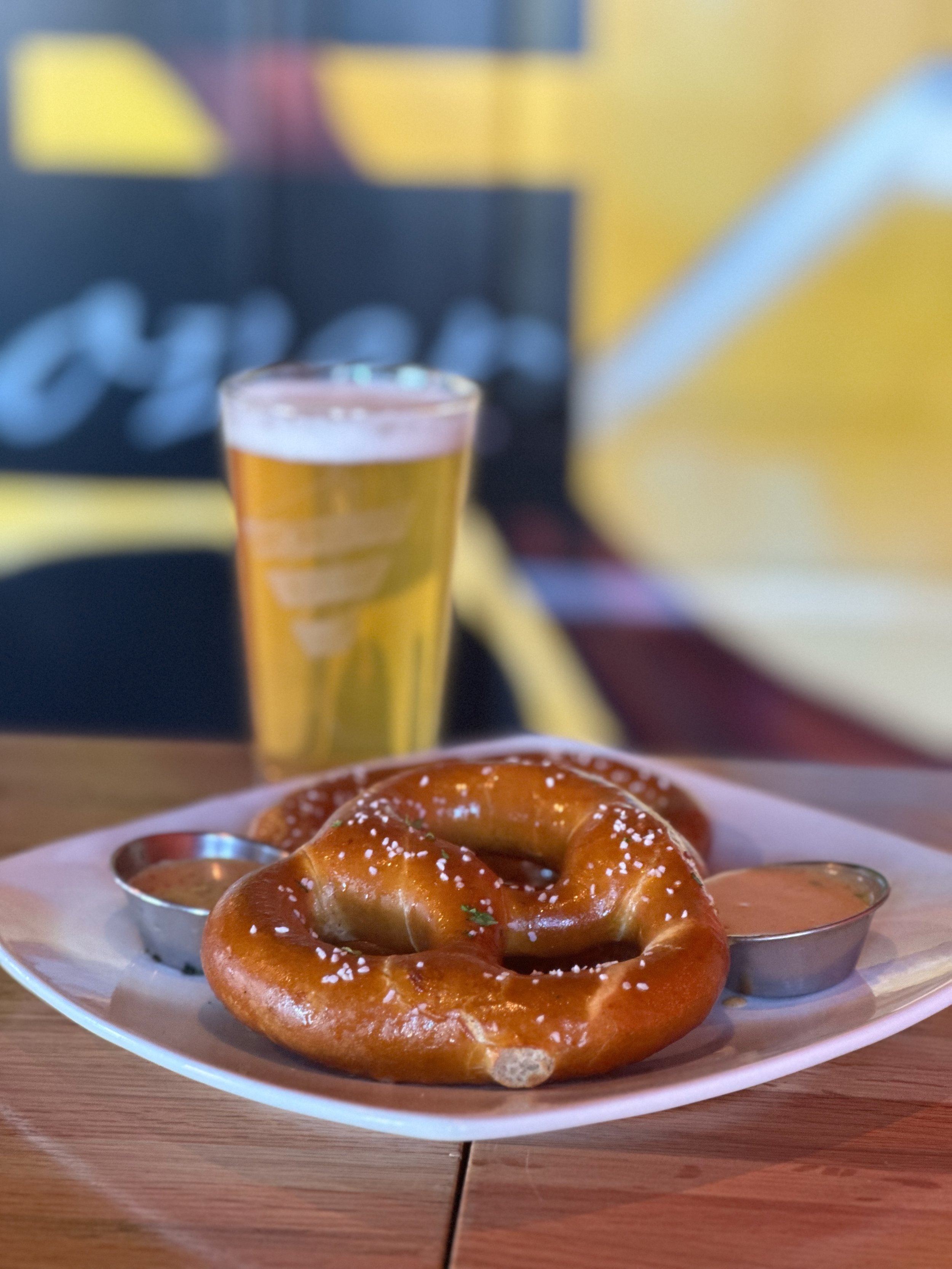 A plate with two large pretzels sprinkled with coarse salt, served with two small cups of dipping sauces, and a glass of beer in the background.