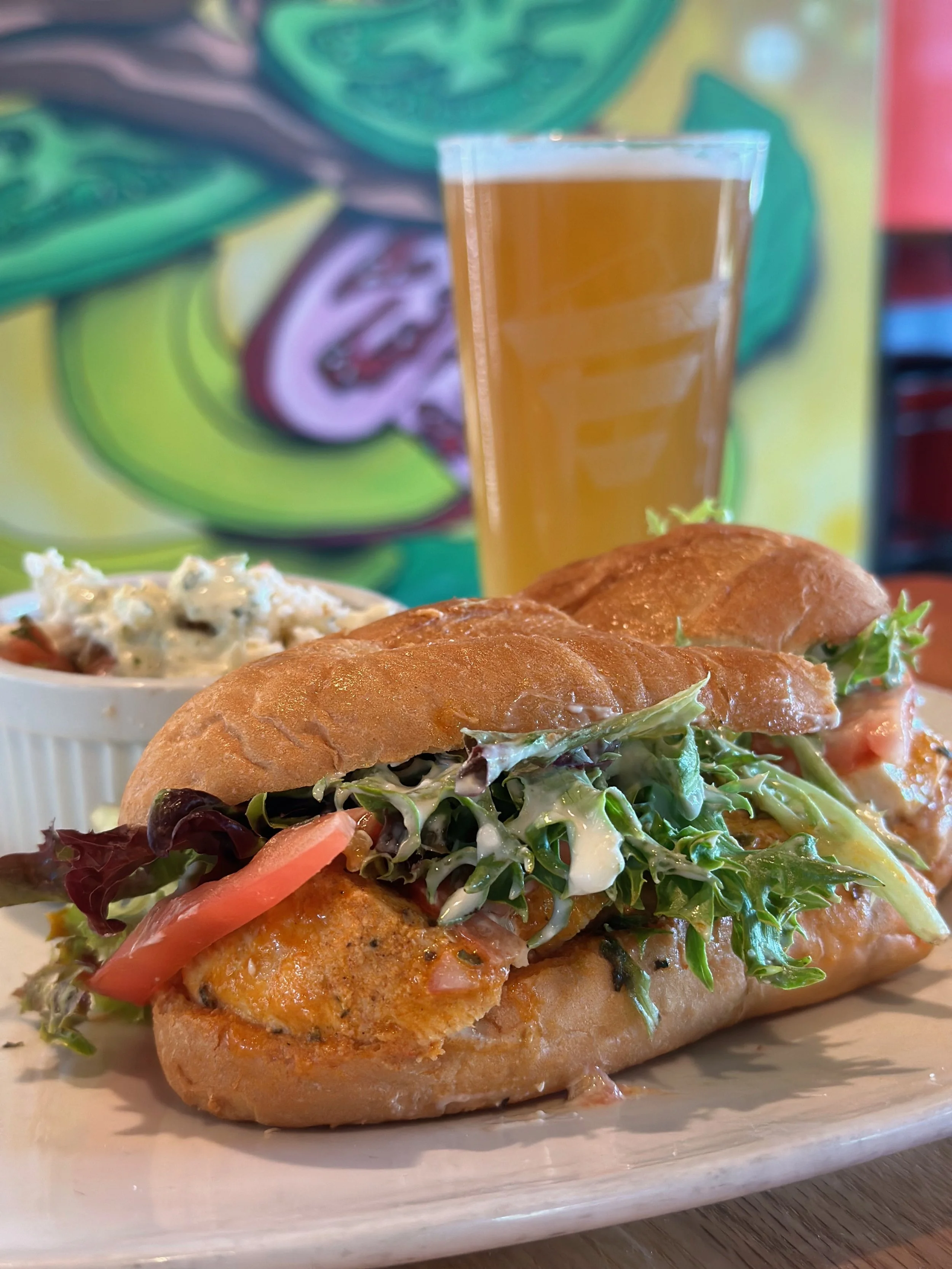 Sandwich with greens, tomato, and chicken sandwich bread, served with a side of potato salad, a glass of beer, and colorful background.
