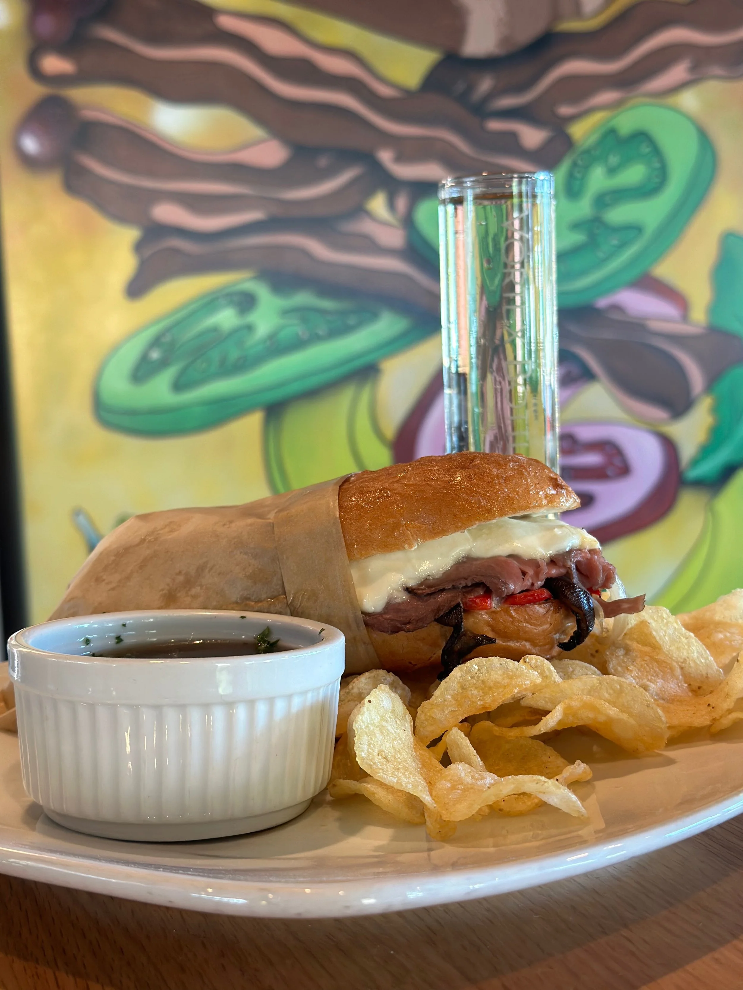 Sandwich with beef, cheese, and vegetables served with potato chips, a small dish of dipping sauce, and a plastic drink stirrer, with colorful abstract mural in background.