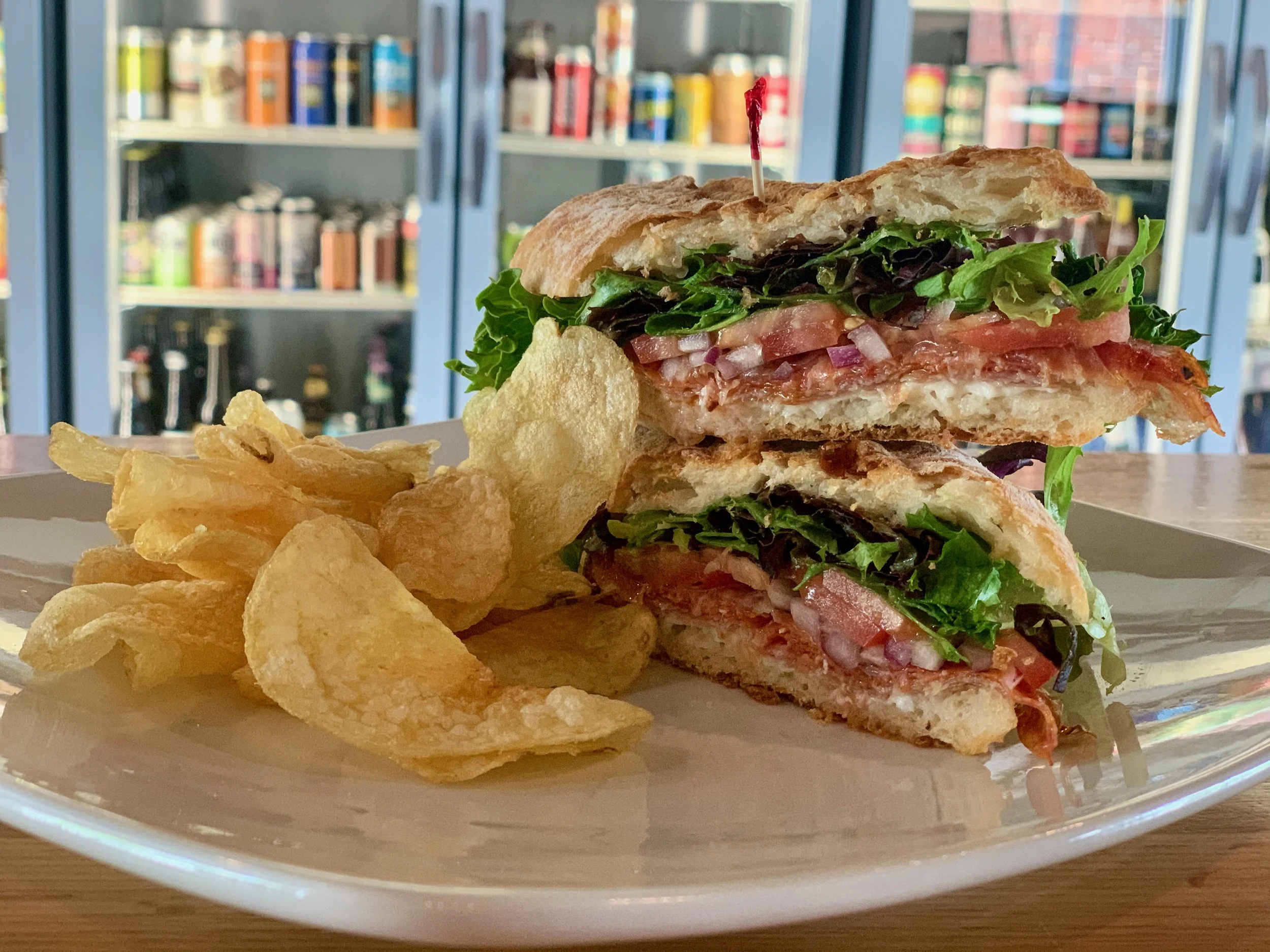 A sandwich with lettuce, tomato, bacon, and spread on toasted bread, served with potato chips on a white plate in a restaurant or cafe setting.