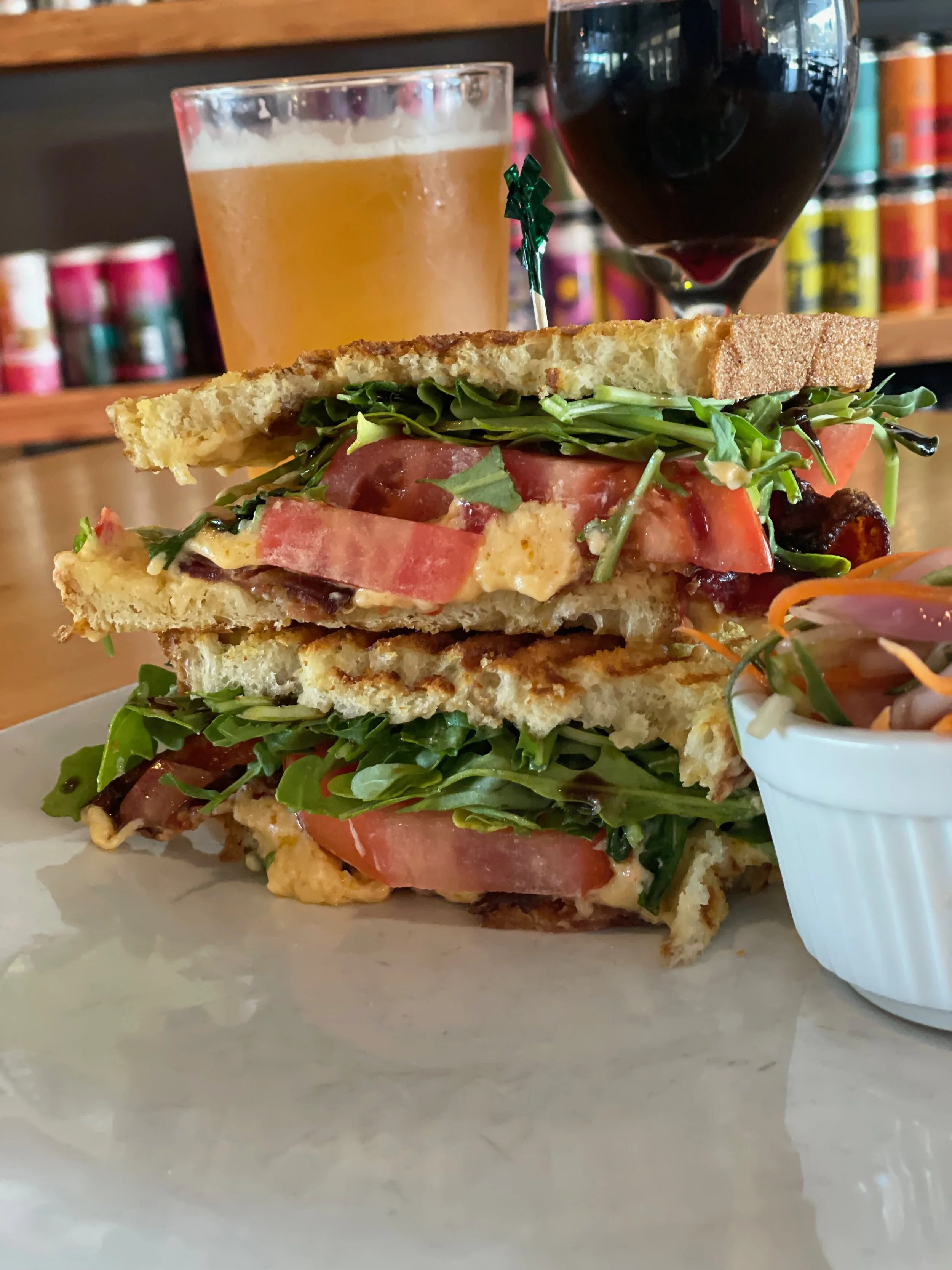 A sandwich with toasted bread, fresh greens, sliced tomatoes, and possibly egg or cheese, served with a side salad and drinks including beer and red wine in a restaurant setting.