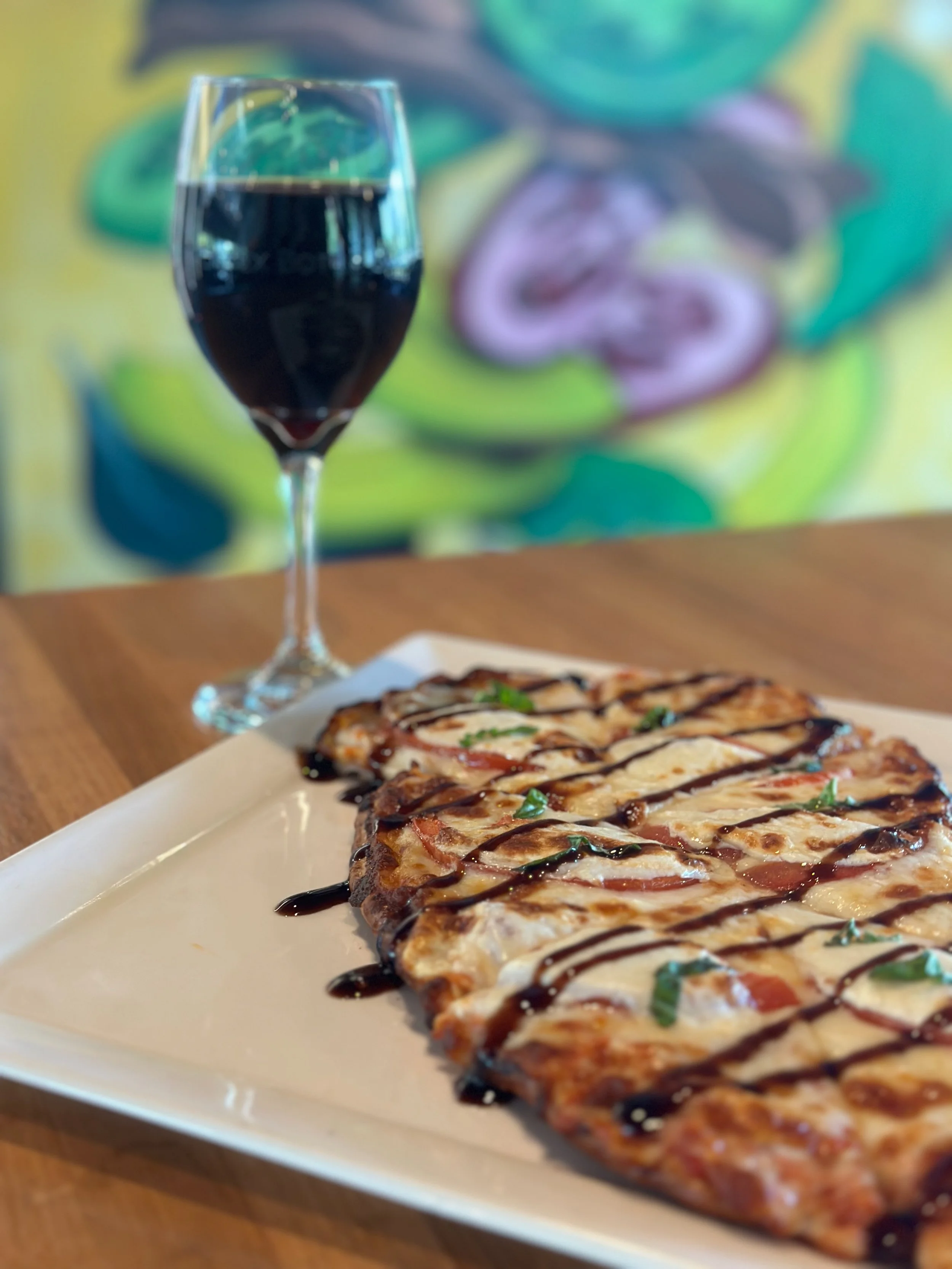 A slice of pizza with drizzled balsamic glaze and chopped basil leaves, a glass of red wine, on a wooden table against a colorful abstract background.