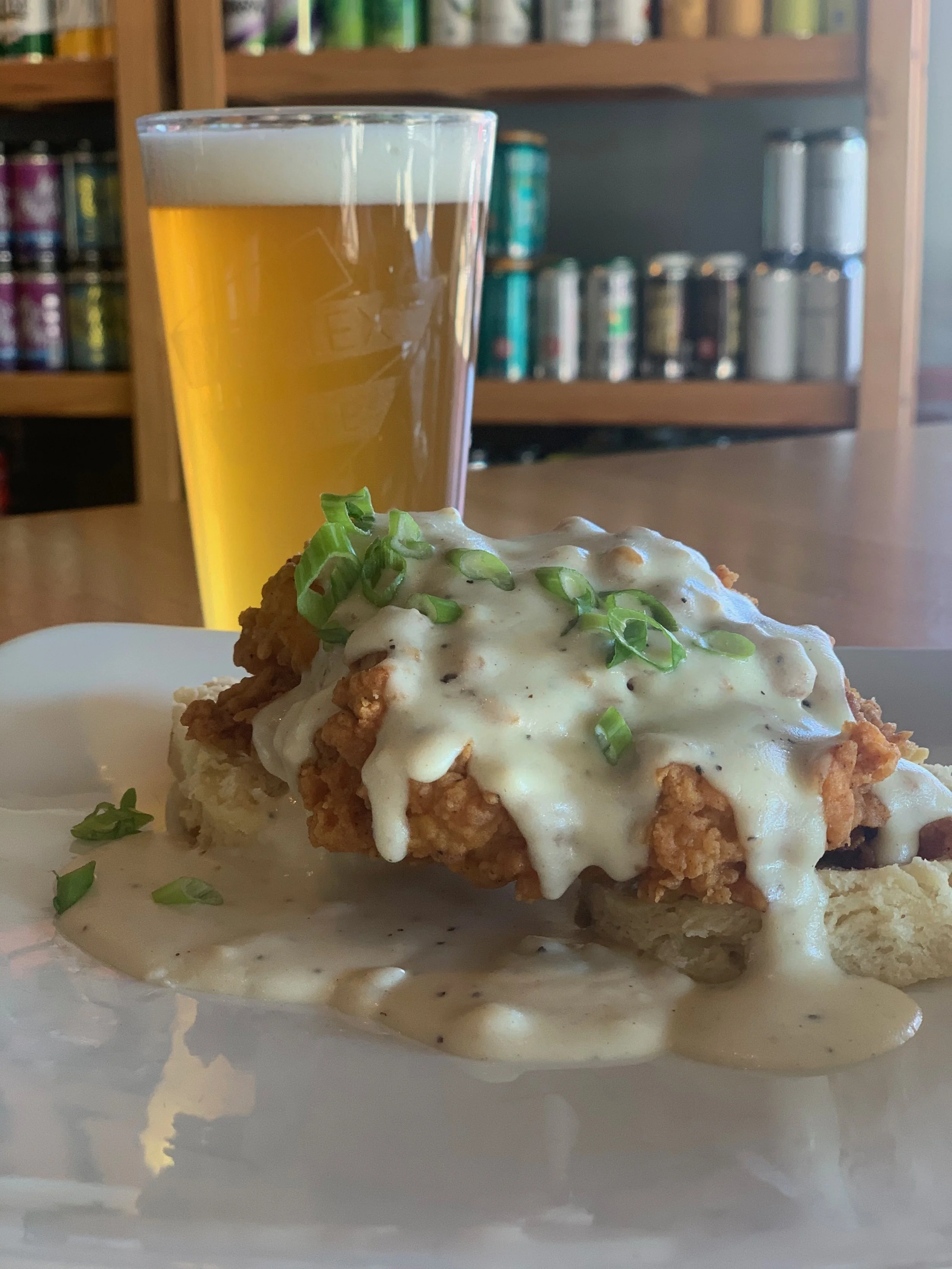 Fried chicken with gravy, garnished with green onions, served on a plate with mashed potatoes, with a glass of beer in the background.