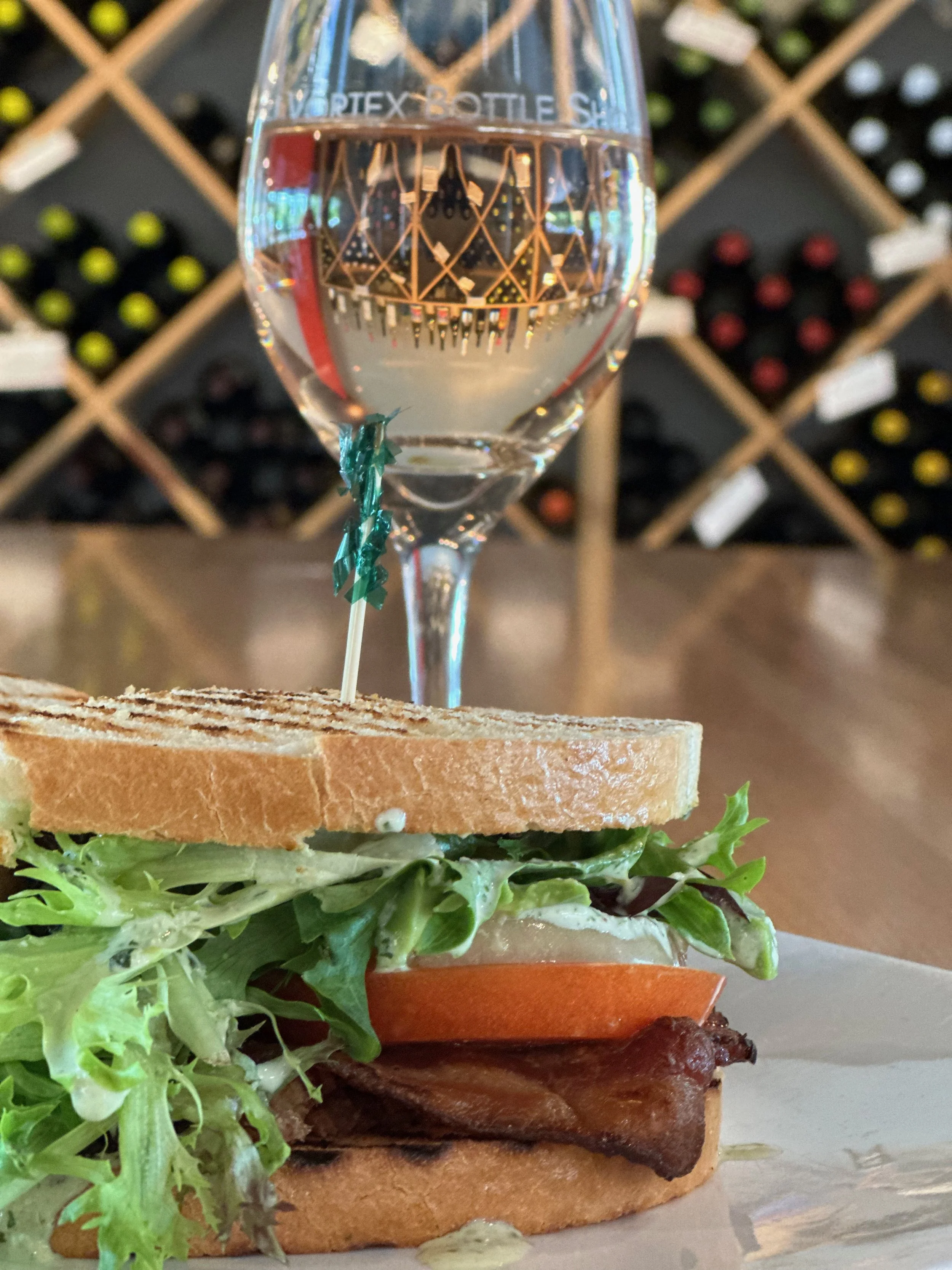 A sandwich with lettuce, tomato, bacon, and creamy sauce on toasted bread, served with a glass of water on a wooden table in a wine cellar.