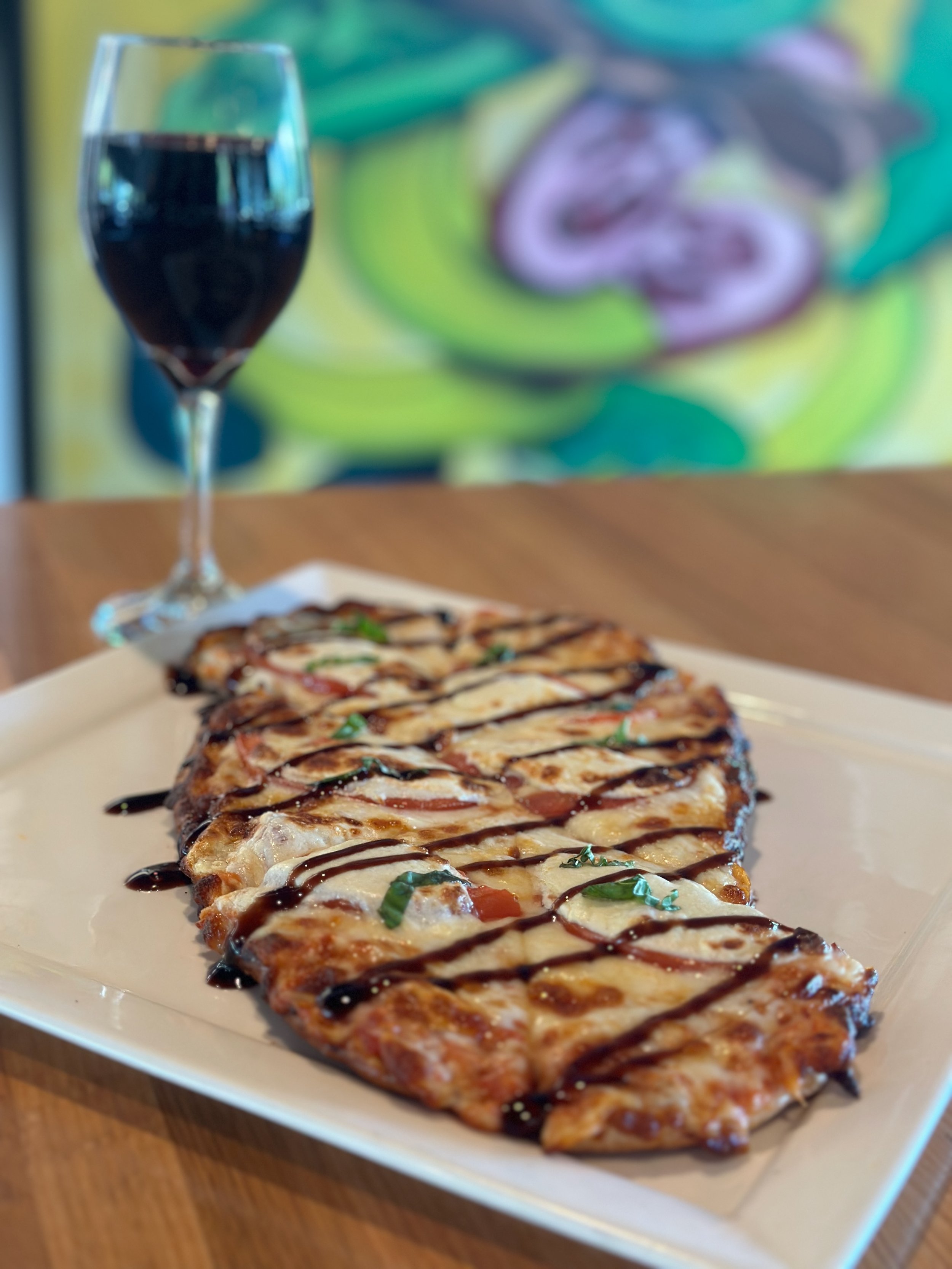 A plate of pizza drizzled with balsamic glaze and chopped herbs, next to a glass of red wine, with a colorful abstract painting in the background.