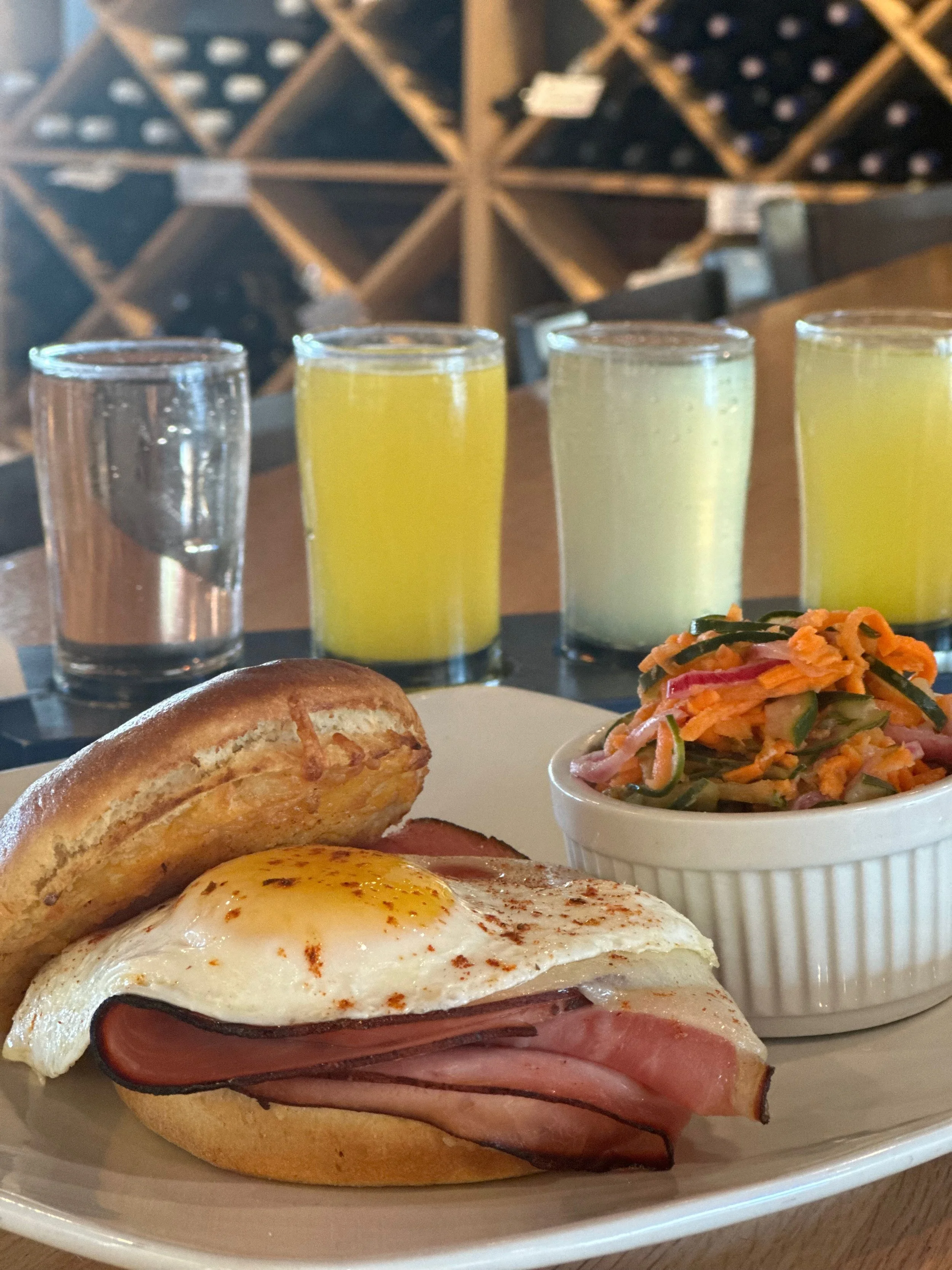 Breakfast sandwich with ham, fried egg, and toasted bun served with a side of coleslaw, four glasses of juice, and water, in a restaurant setting with wine bottles in the background.