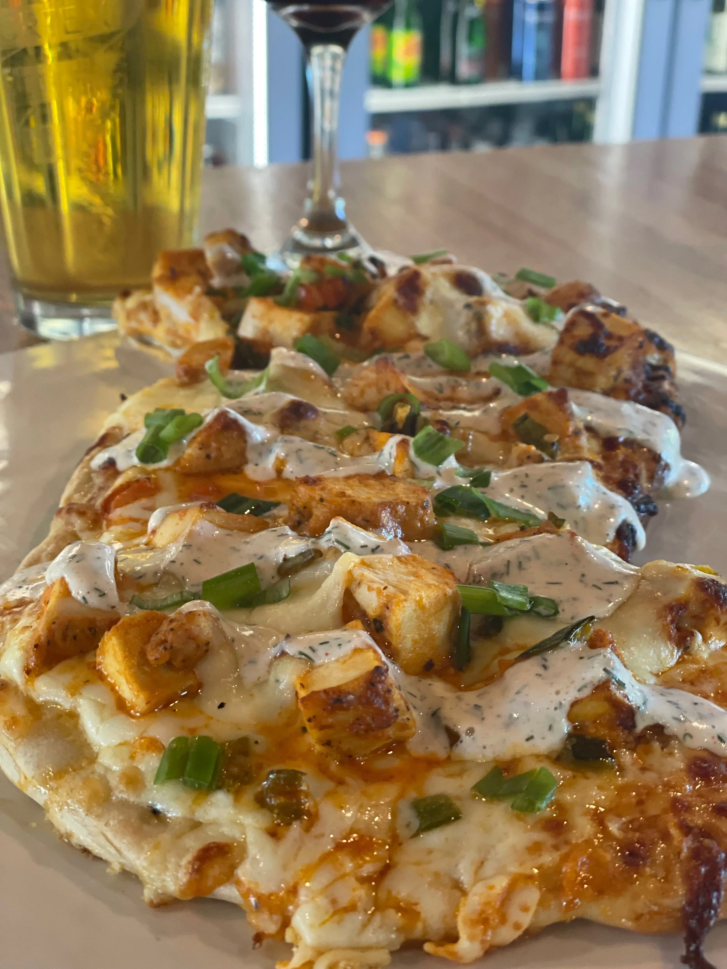 Close-up of a slice of pizza topped with grilled chicken pieces, green onions, and creamy dressing on a white plate, with a glass of beer and a wine glass in the background.