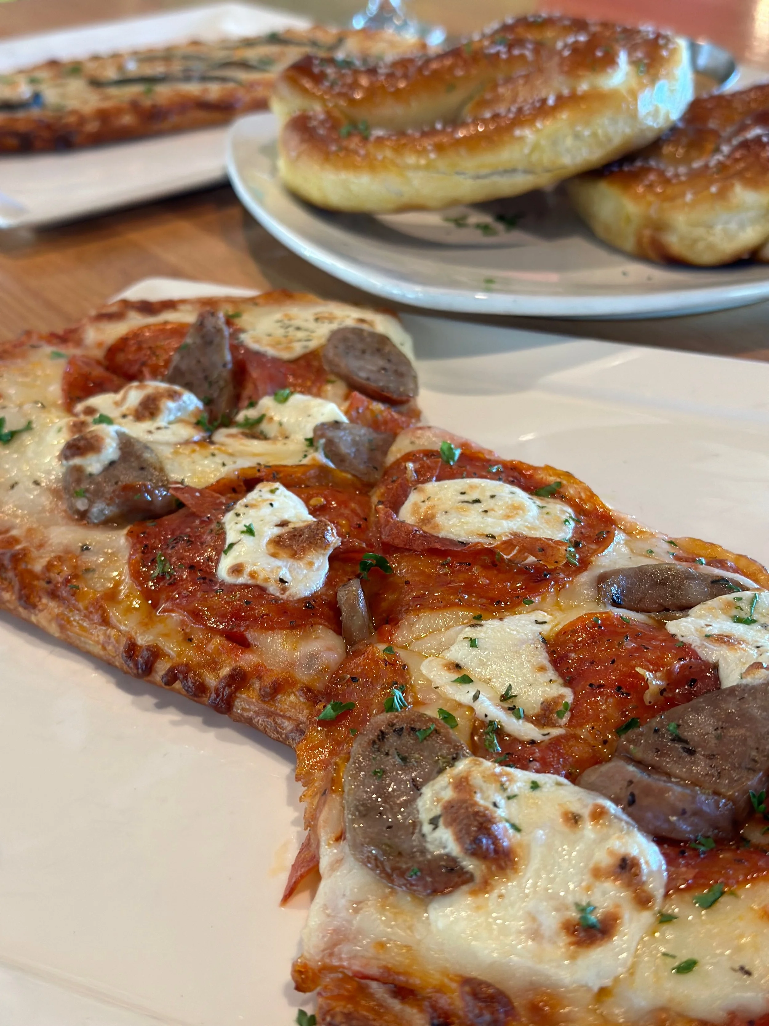 A slice of pepperoni and sausage pizza with melted cheese, served on a white plate, with a folded calzone and a grilled bread in the background.