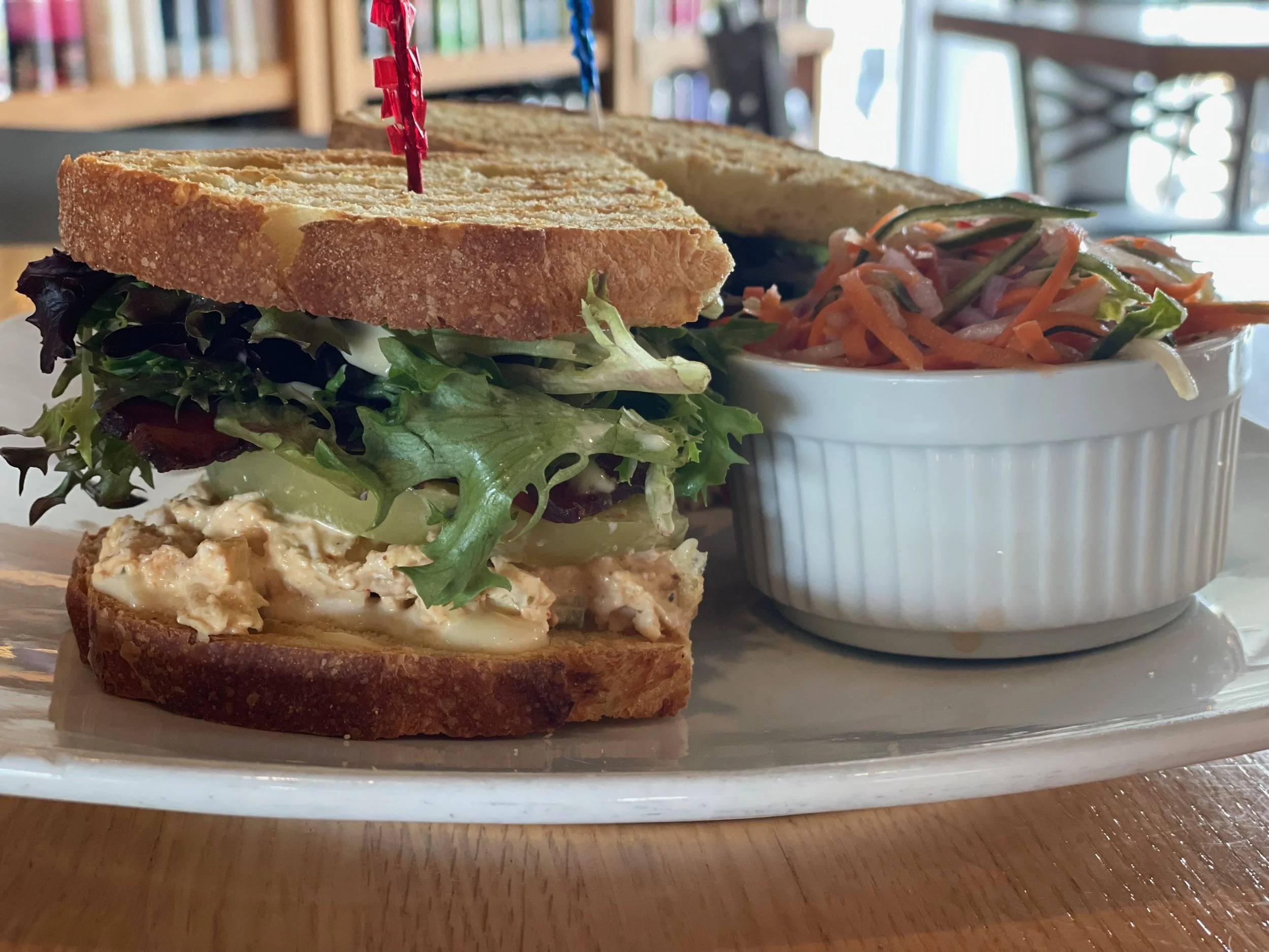A close-up of a sandwich with leaf lettuce, cucumbers, and chicken salad on toasted bread, served on a white plate with sides of coleslaw in a white bowl, set on a wooden table in a cafe environment.