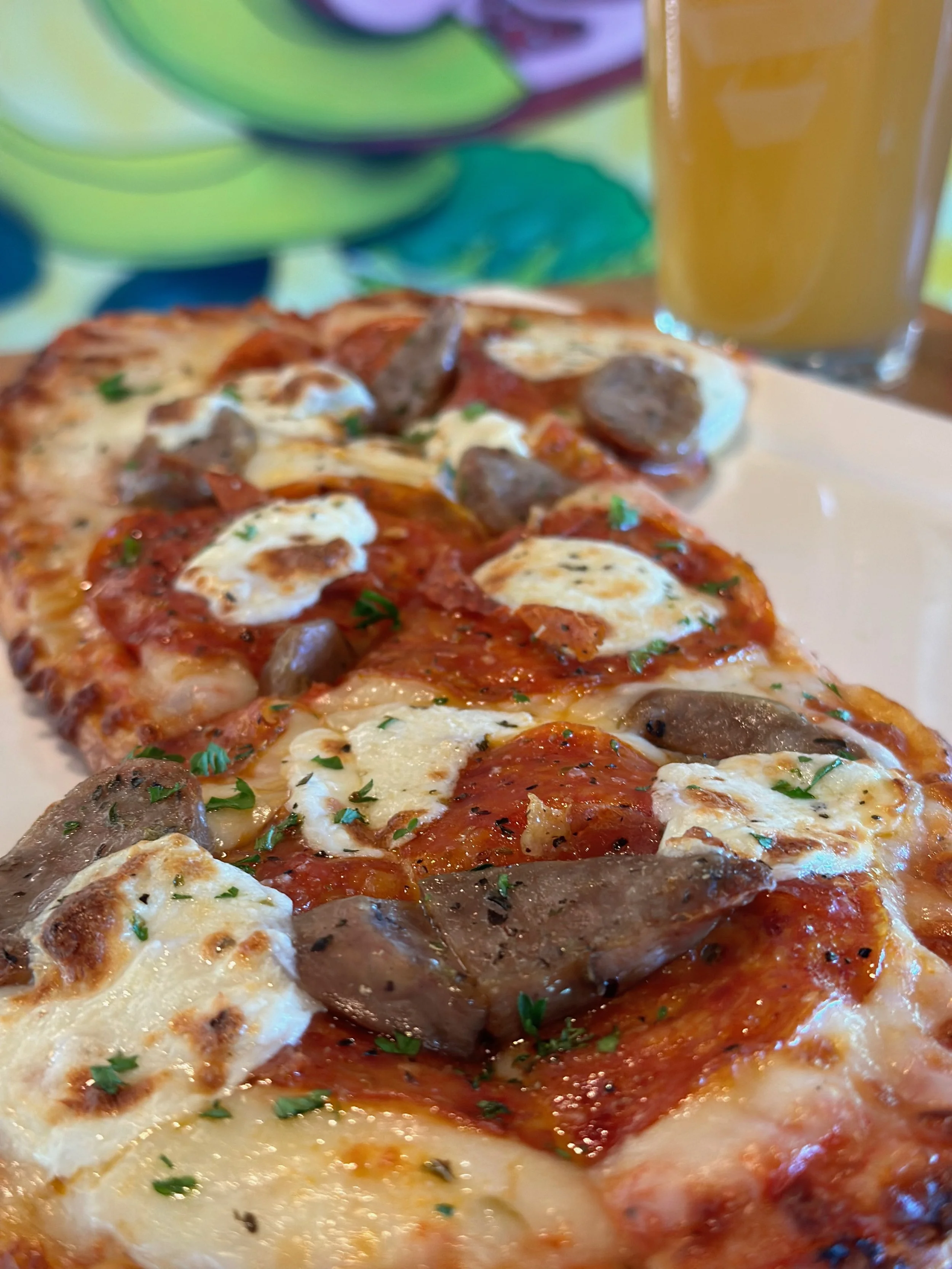 Close-up of a pepperoni and sausage pizza with melted cheese and herbs, with a glass of beer in the background.