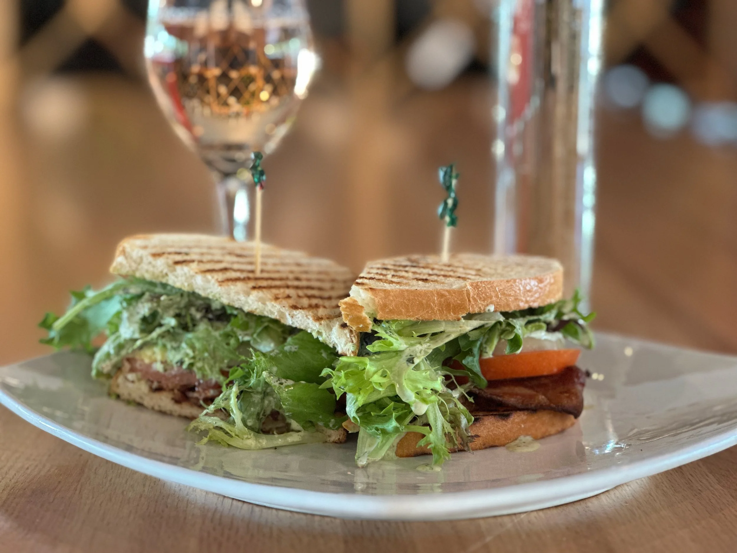 Half of a grilled sandwich with lettuce, tomato, and meat on a white plate, with a glass of water and a glass of white wine in the background.