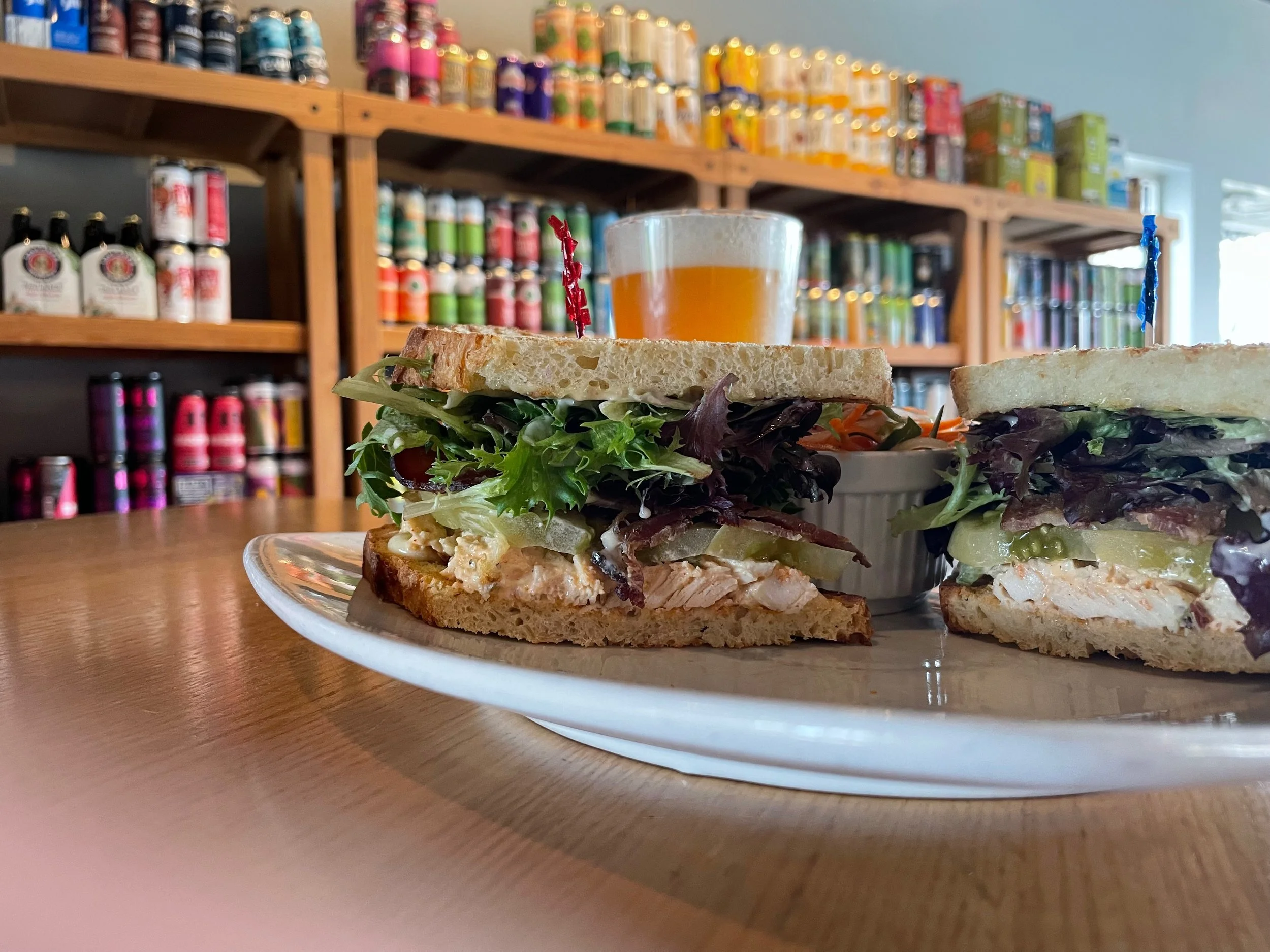 Close-up of two sandwiches on a plate with a glass of beer and coleslaw in the background, on a wooden table in a cozy café or deli, with shelves of canned goods and spices on the wall.