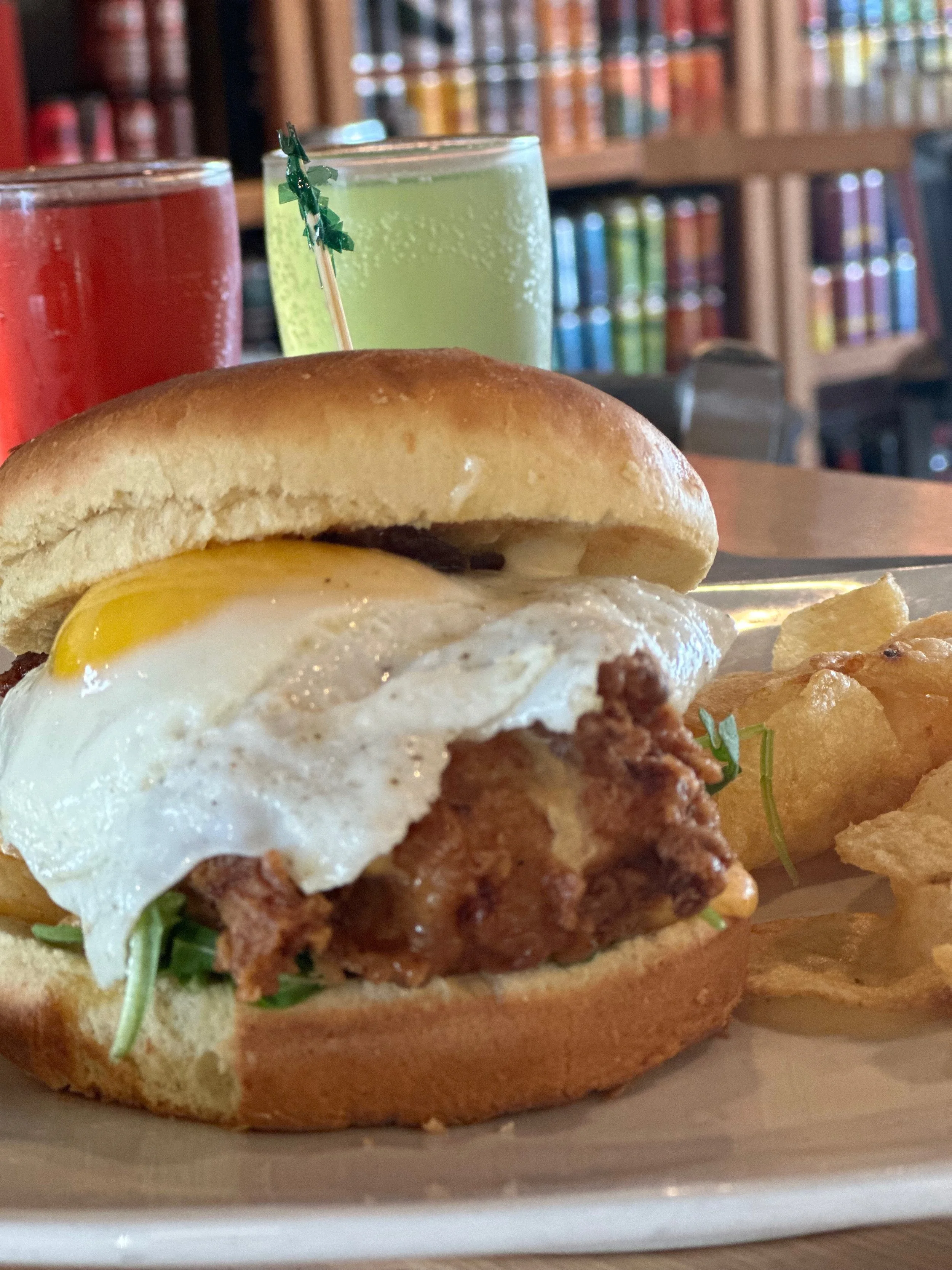 Close-up of a breakfast sandwich with fried chicken, sunny-side-up egg, and greens on a bun, served with potato chips. Behind the sandwich, there are two drinks, one red and one green, with a blurred background of bookshelves.