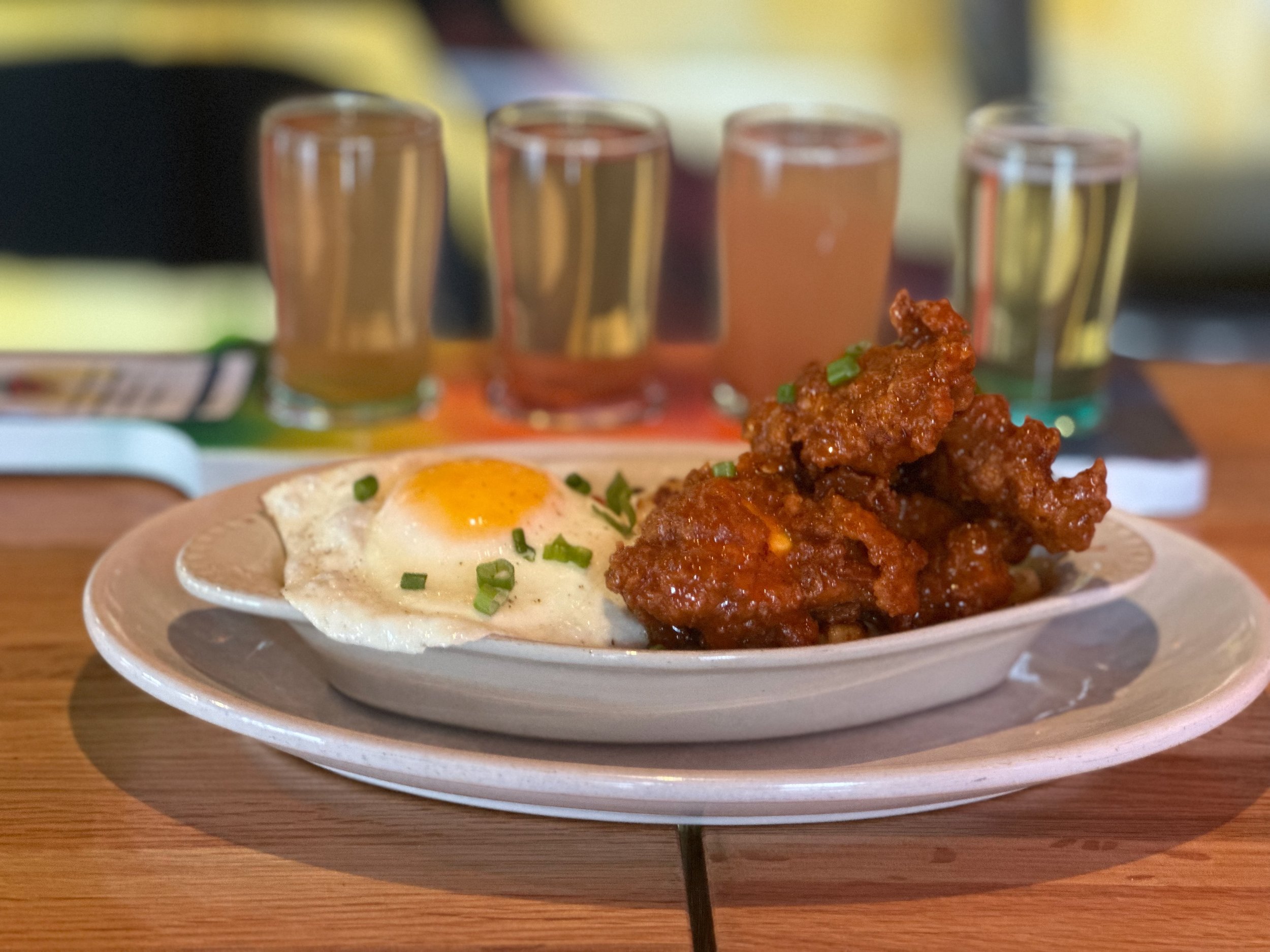 A plate with fried chicken and a fried egg garnished with chopped green onions, with four shot glasses filled with different colored liquids in the background.