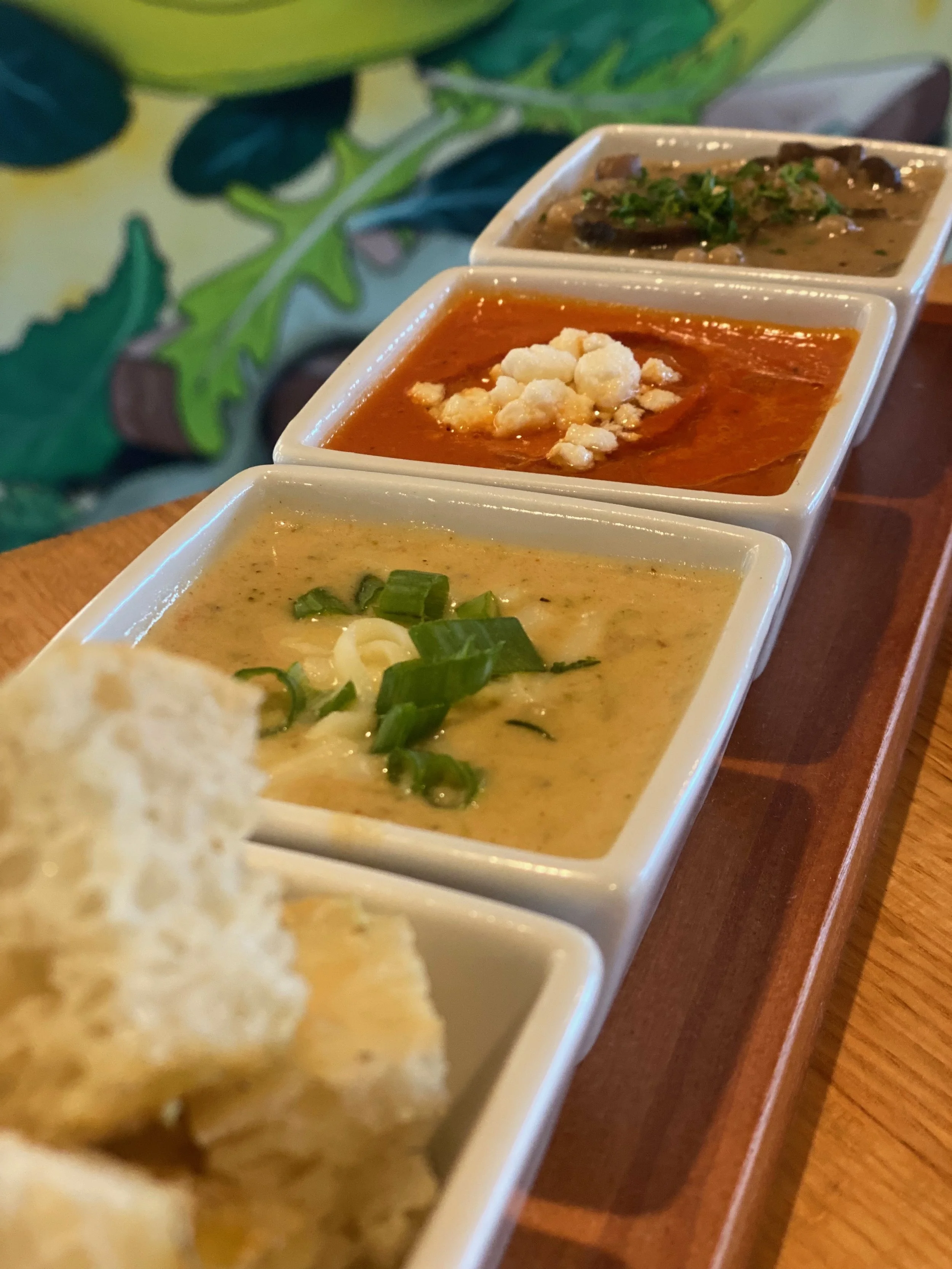 A wooden platter with four small white ramekins containing different types of Indian curries and sauces, garnished with green herbs and cheese, with a piece of bread in the foreground.
