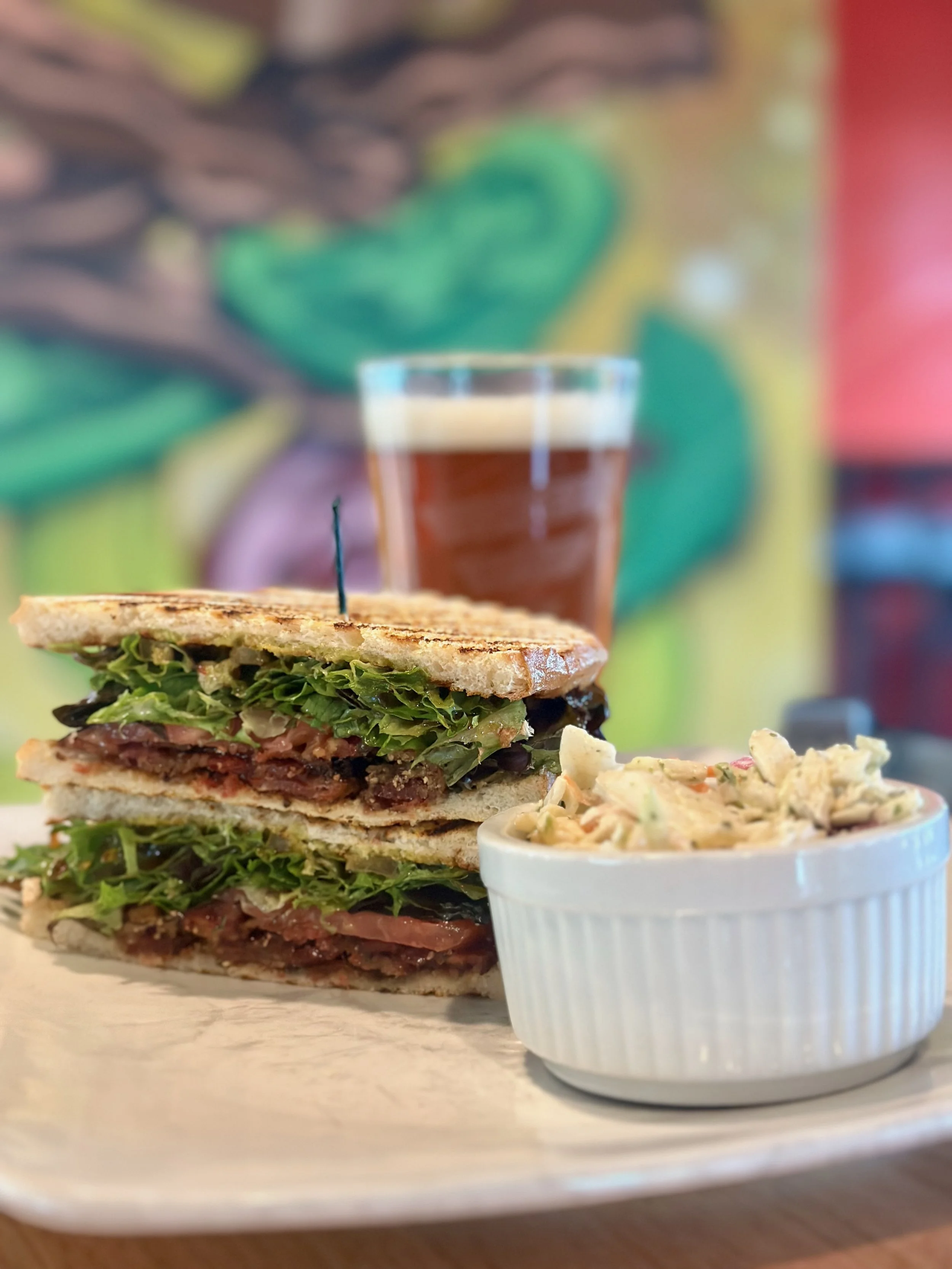 Close-up of a toasted sandwich with layers of lettuce, tomato, and meat, served with a side of creamy coleslaw and a glass of iced tea in the background, with a colorful abstract mural in the background.