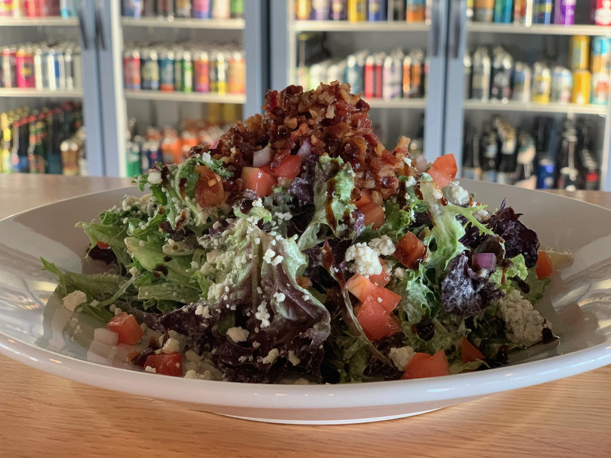 A plate of salad with lettuce, tomatoes, feta cheese, bacon bits, and a balsamic glaze on top, placed on a wooden table with a bookshelf in the background.