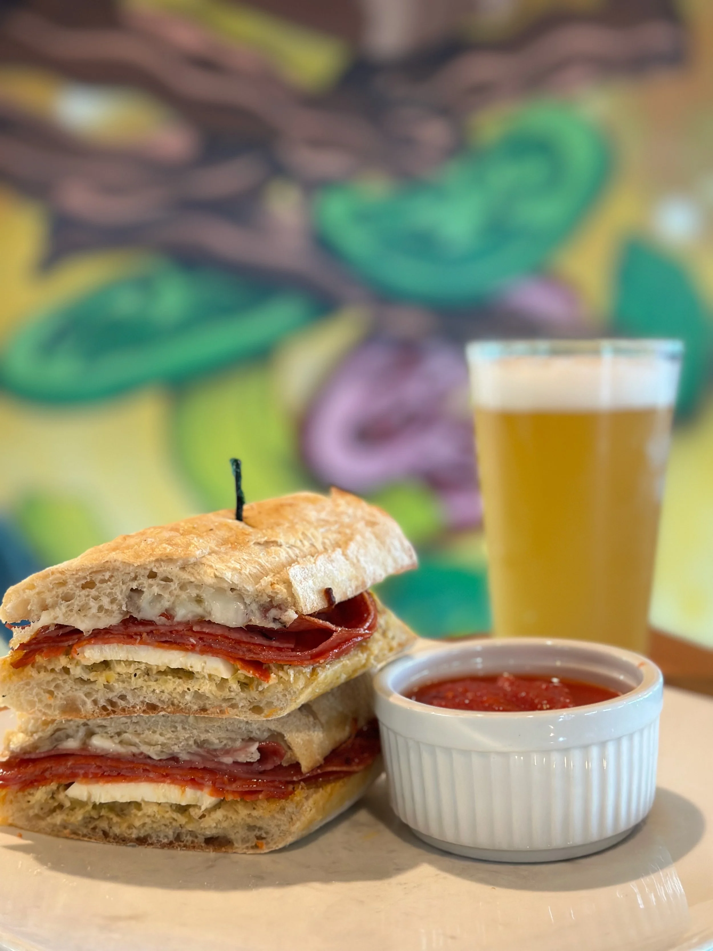 A sandwich with slices of meat and cheese on a crusty bread roll, served with a small bowl of marinara sauce and a glass of beer in the background.