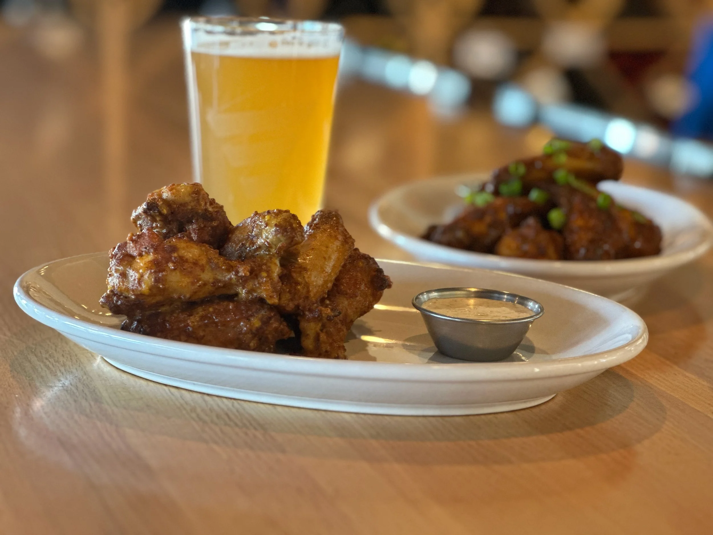 Fried chicken wings with dipping sauce, a glass of beer, and a plate of chicken wings with green onions in the background, set on a wooden table.