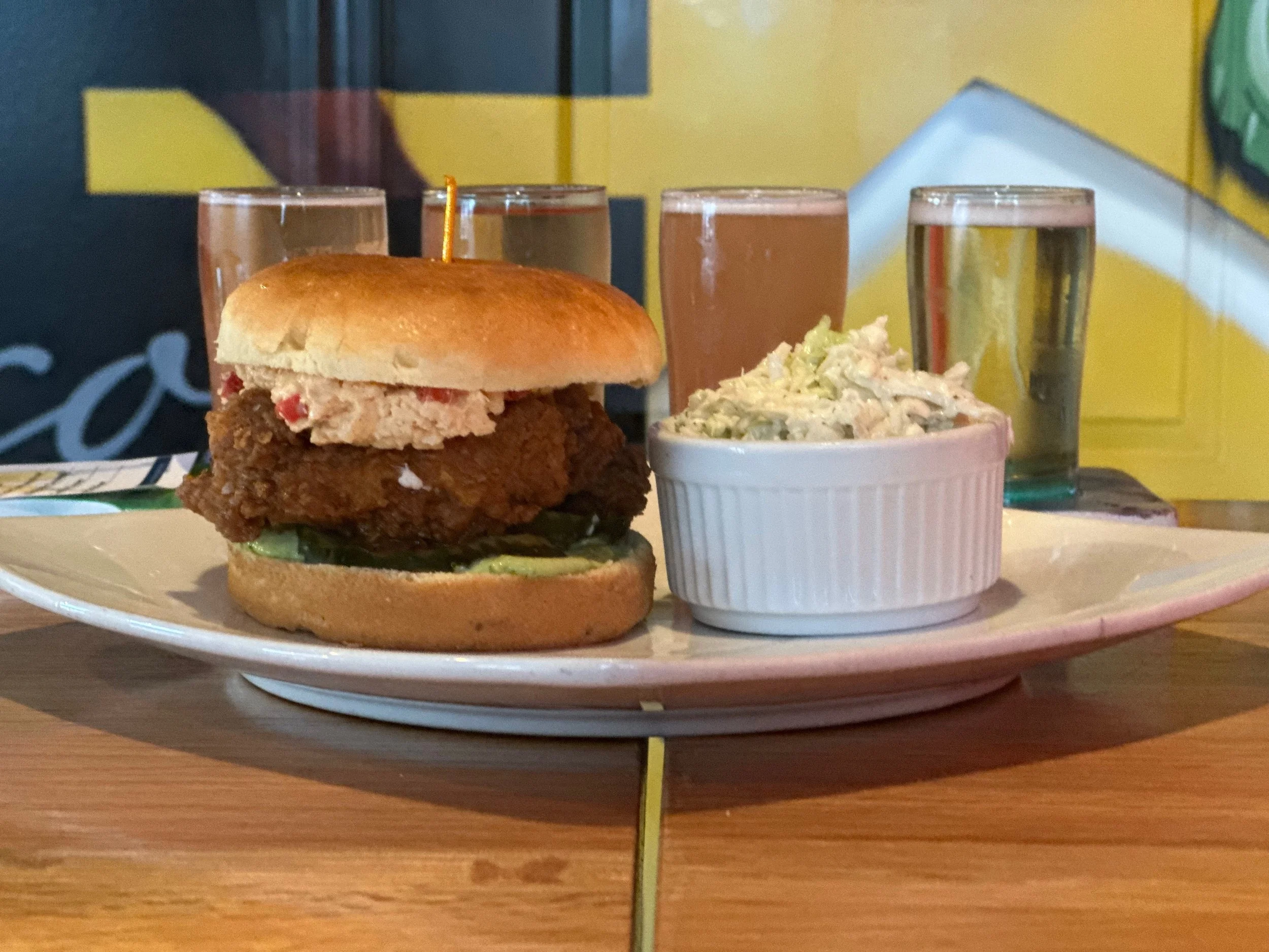 Fried chicken sandwich with lettuce and a topping on a bun, alongside a bowl of coleslaw and four glasses of varying drinks, on a white plate on a wooden table.