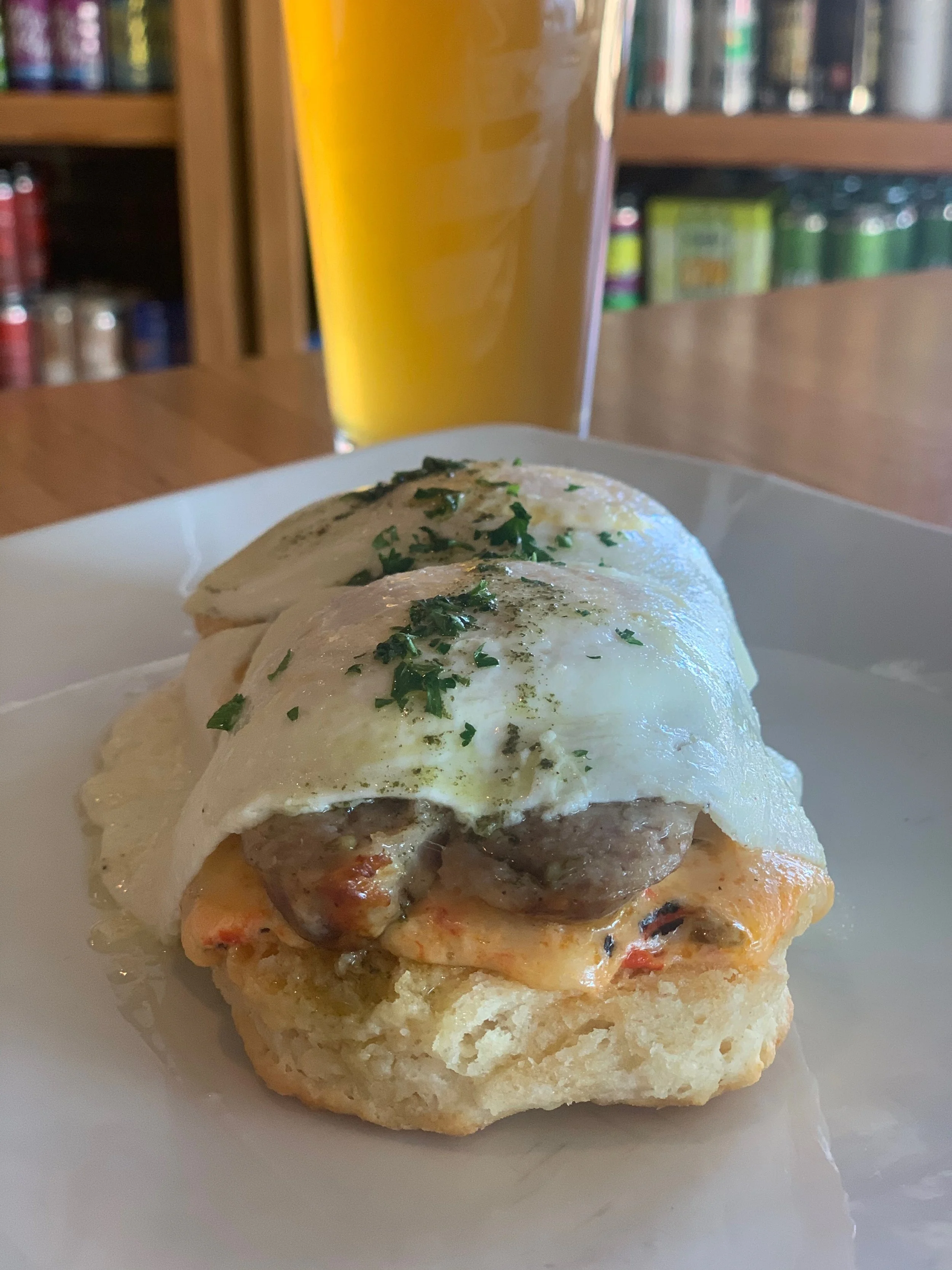Close-up of an open-faced breakfast biscuit topped with fried eggs, mushrooms, cheese, and parsley with a glass of beer in the background.