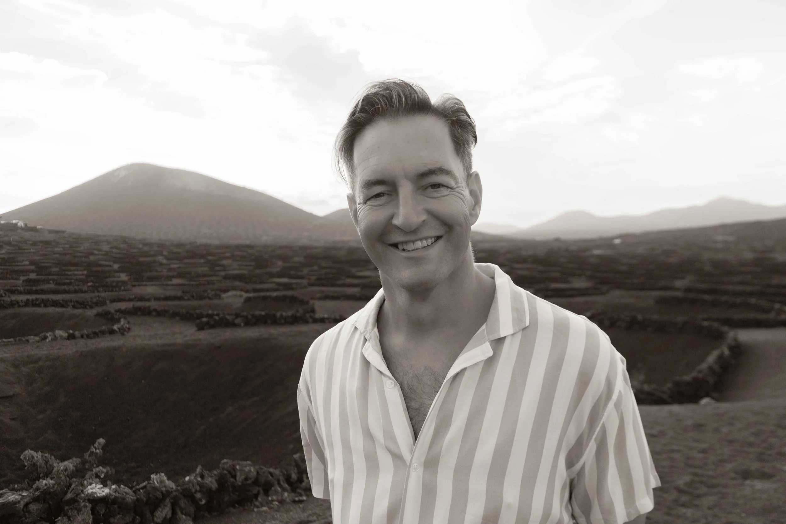 A smiling man in a striped shirt standing outdoors with a mountain and farmland in the background.