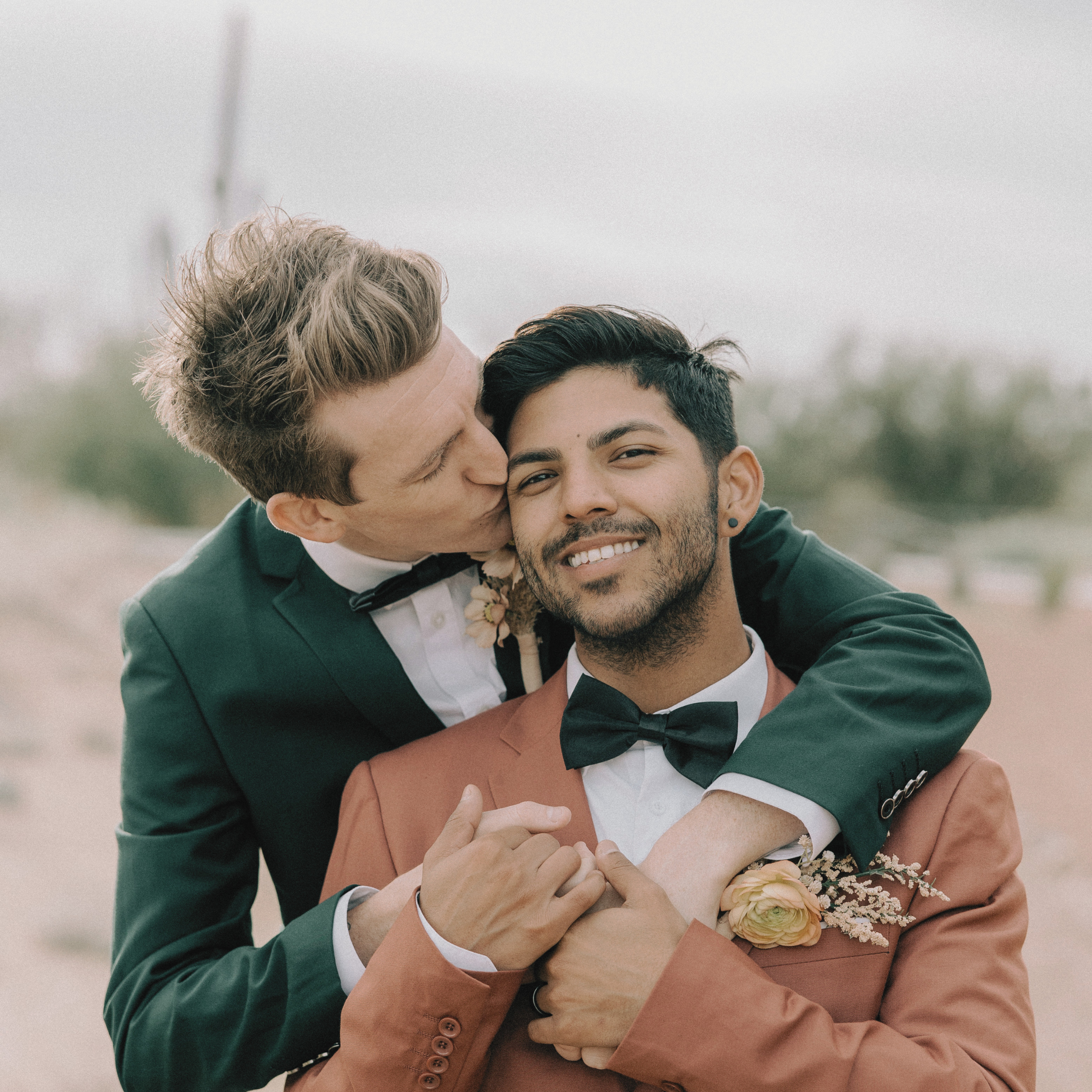 Two men dressed in tuxedos, one with blonde curly hair and the other with dark hair and beard, sharing a kiss on the cheek at a wedding or celebration.