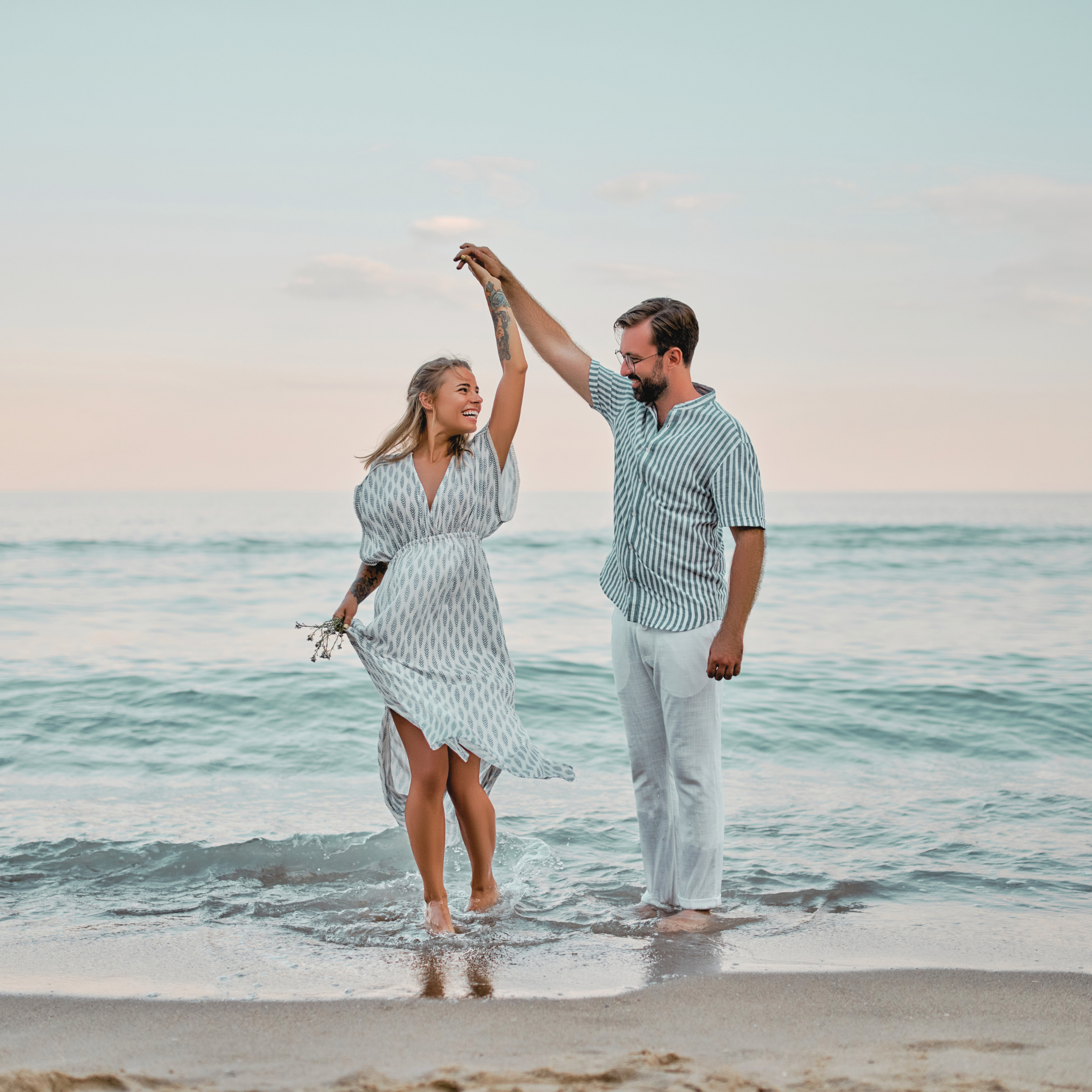 A smiling woman in a summer dress with tattoos on her arm raising her hand in a high-five with a man in a striped shirt at the beach, standing in the water near the shore during sunset.