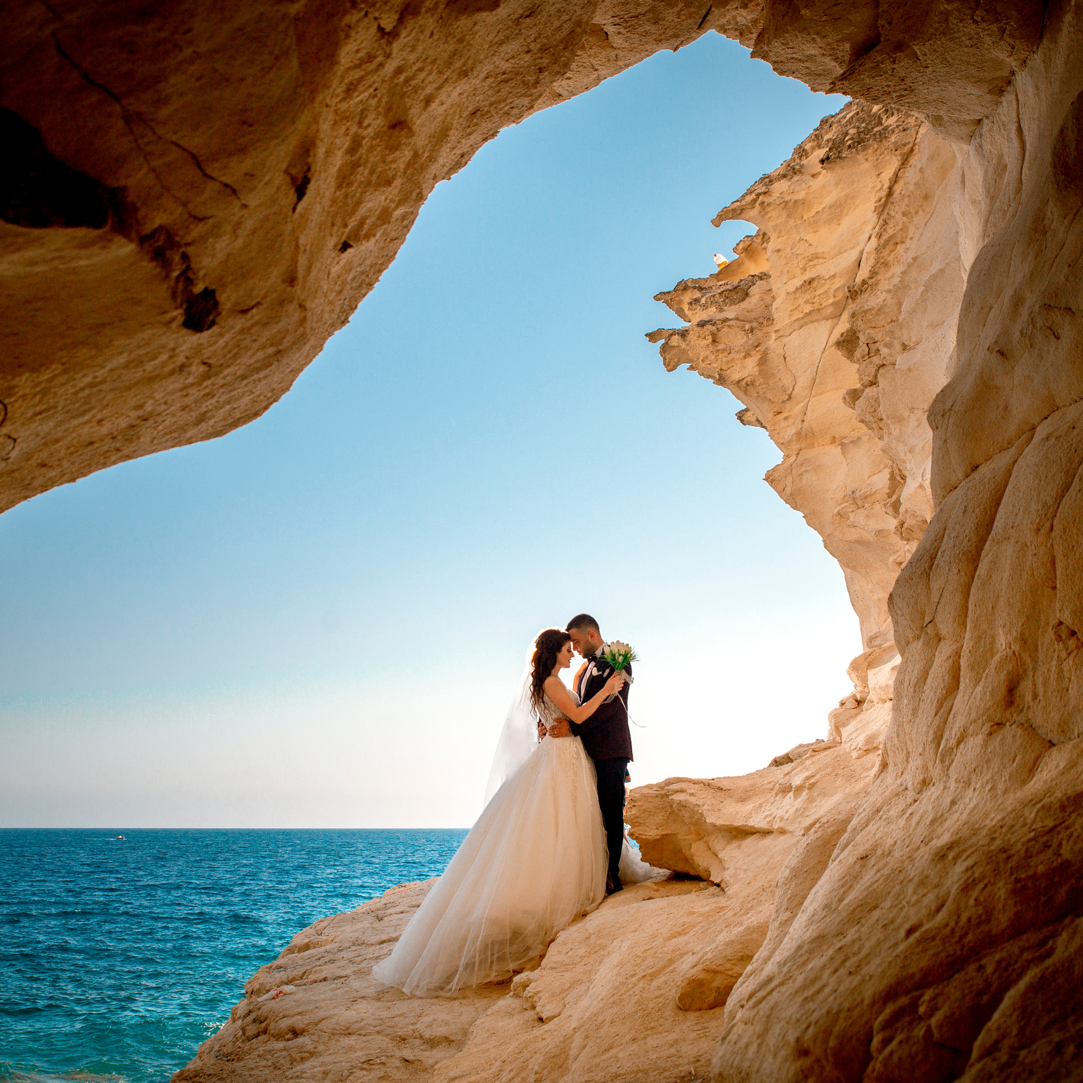 A bride and groom standing close inside a large natural rock formation by the ocean, with the bride in a white wedding dress and the groom in a black tuxedo, during daytime.