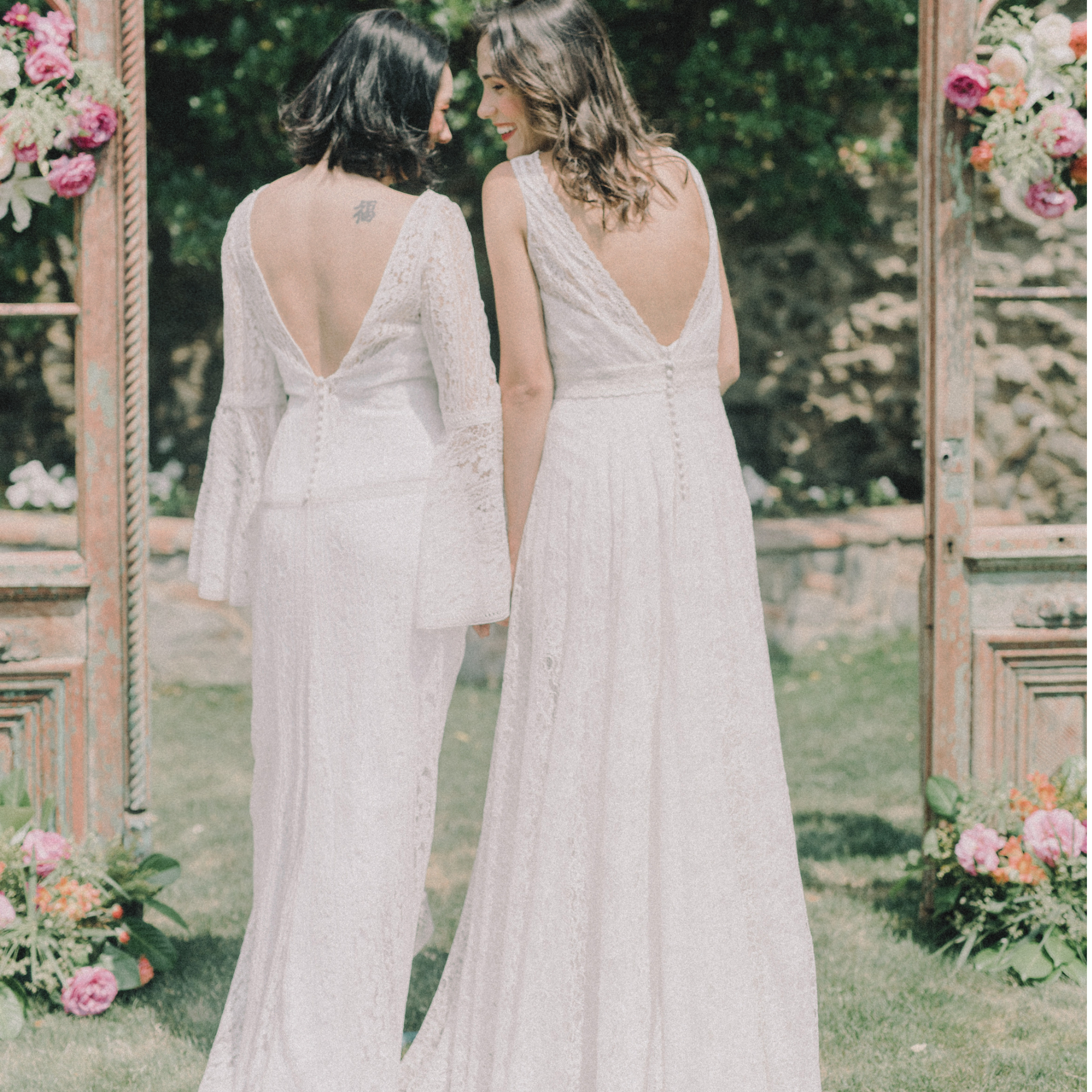 Two women in wedding dresses standing close together outdoors, smiling, with a floral archway decorated with pink and white flowers in the background.